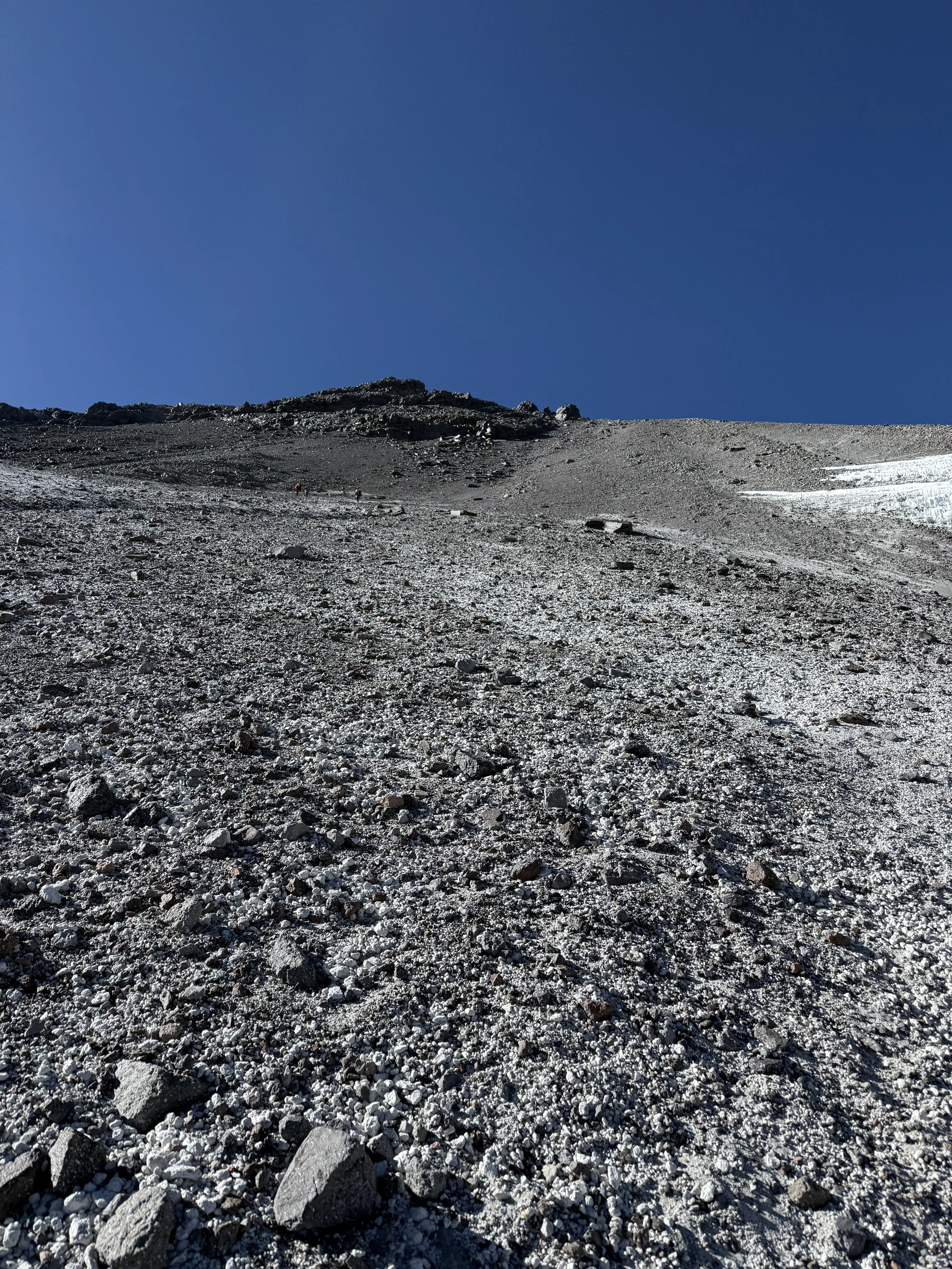 Steep, rocky mountain slope with sparse snow patches, under a clear blue sky.