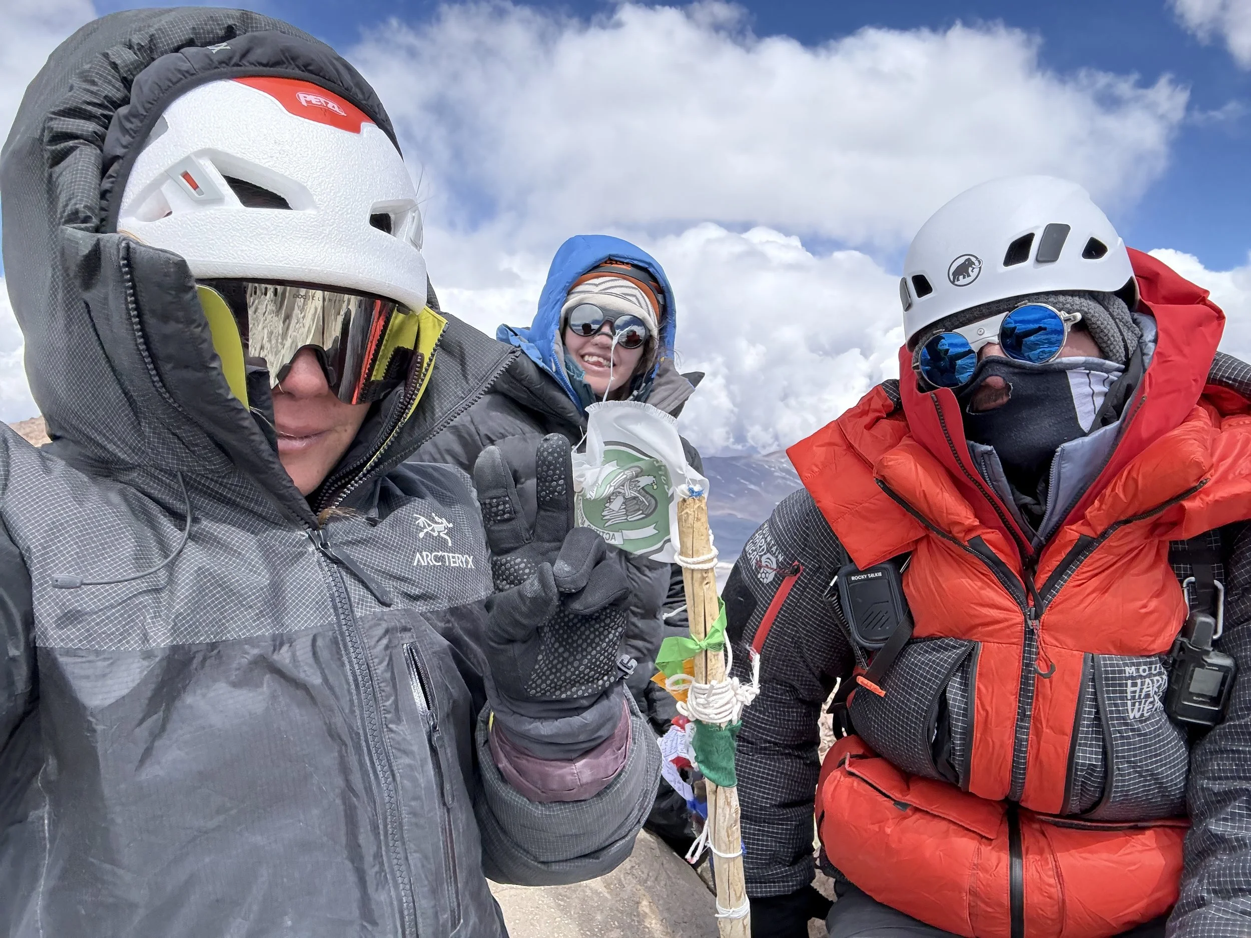 Three climbers in winter gear on a mountain, with a cloudy sky in the background. One person holds a trekking pole decorated with ribbons and fabric.