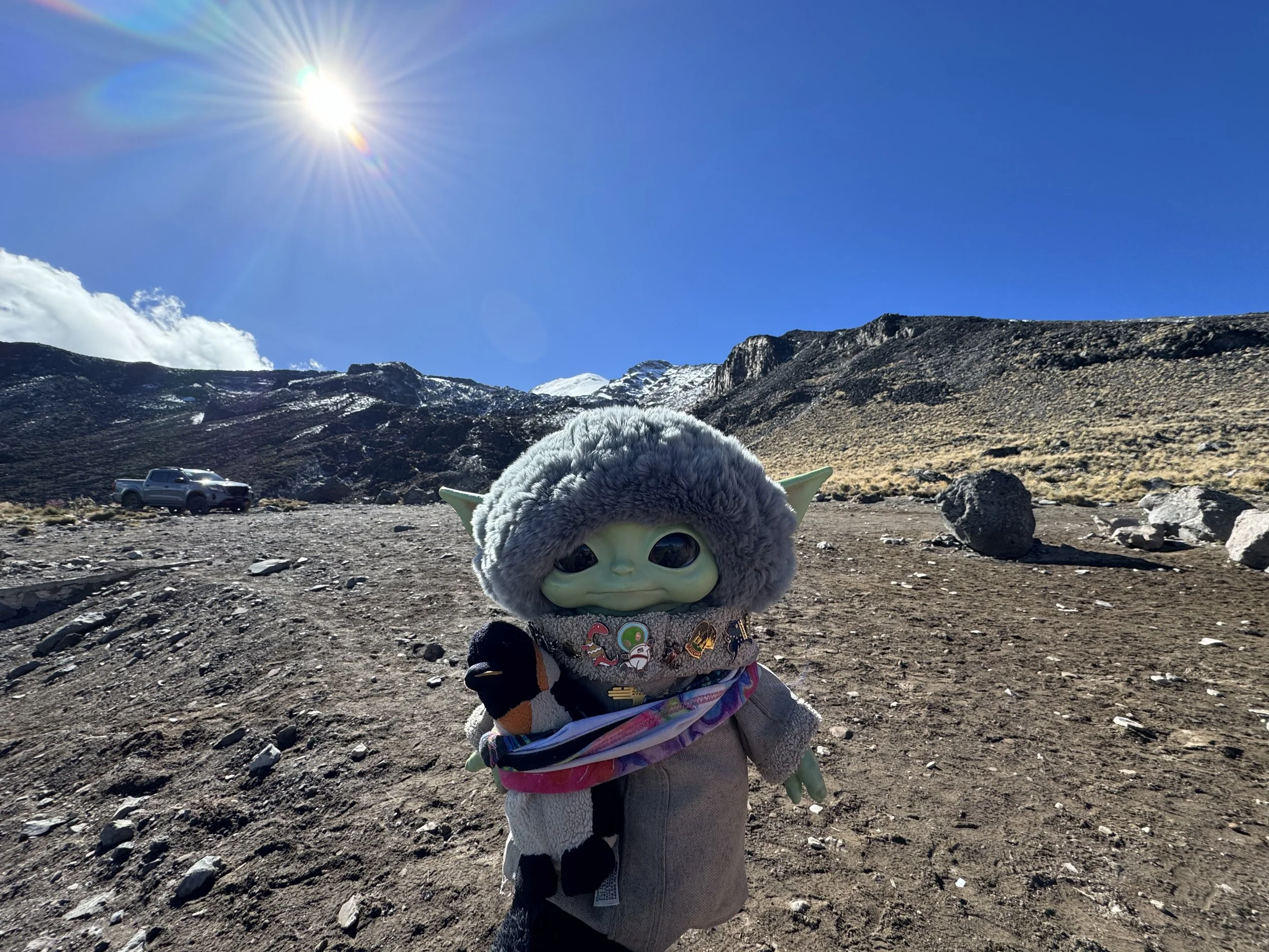 A plush toy resembling Grogu from Star Wars stands in a rocky, mountainous landscape under a bright blue sky with the sun shining.