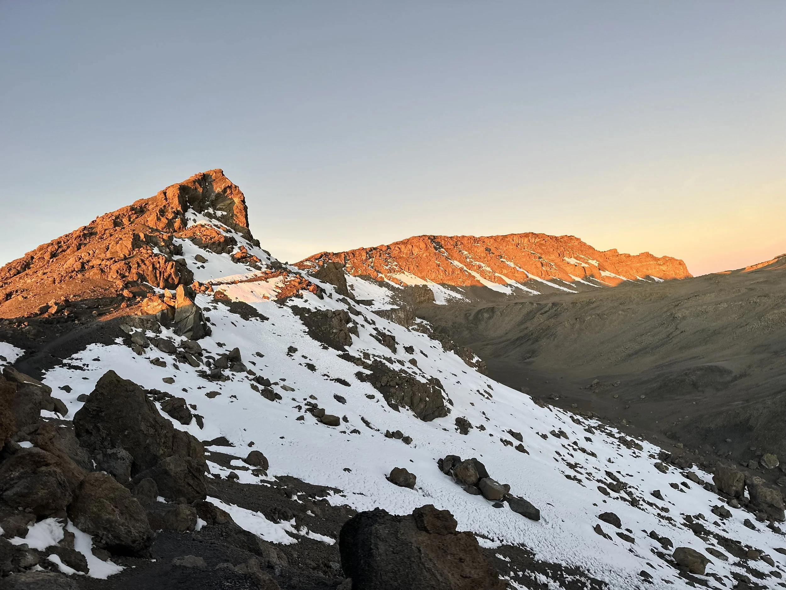 A rugged mountain landscape during sunset with snow patches and rocky terrain.