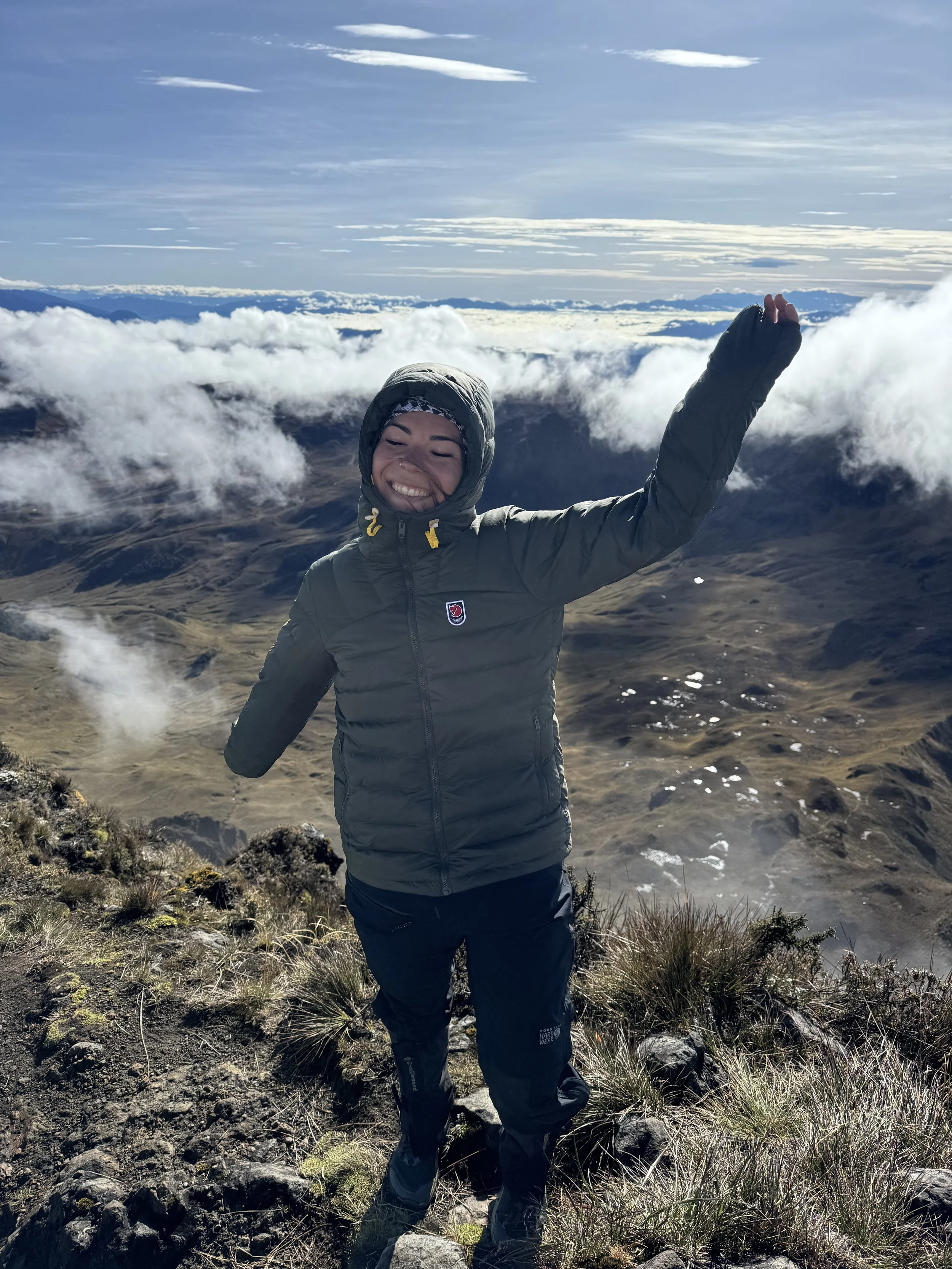 A woman smiling and celebrating on a mountain with her arms raised, wearing a gray jacket and black pants, with clouds and mountains in the background.