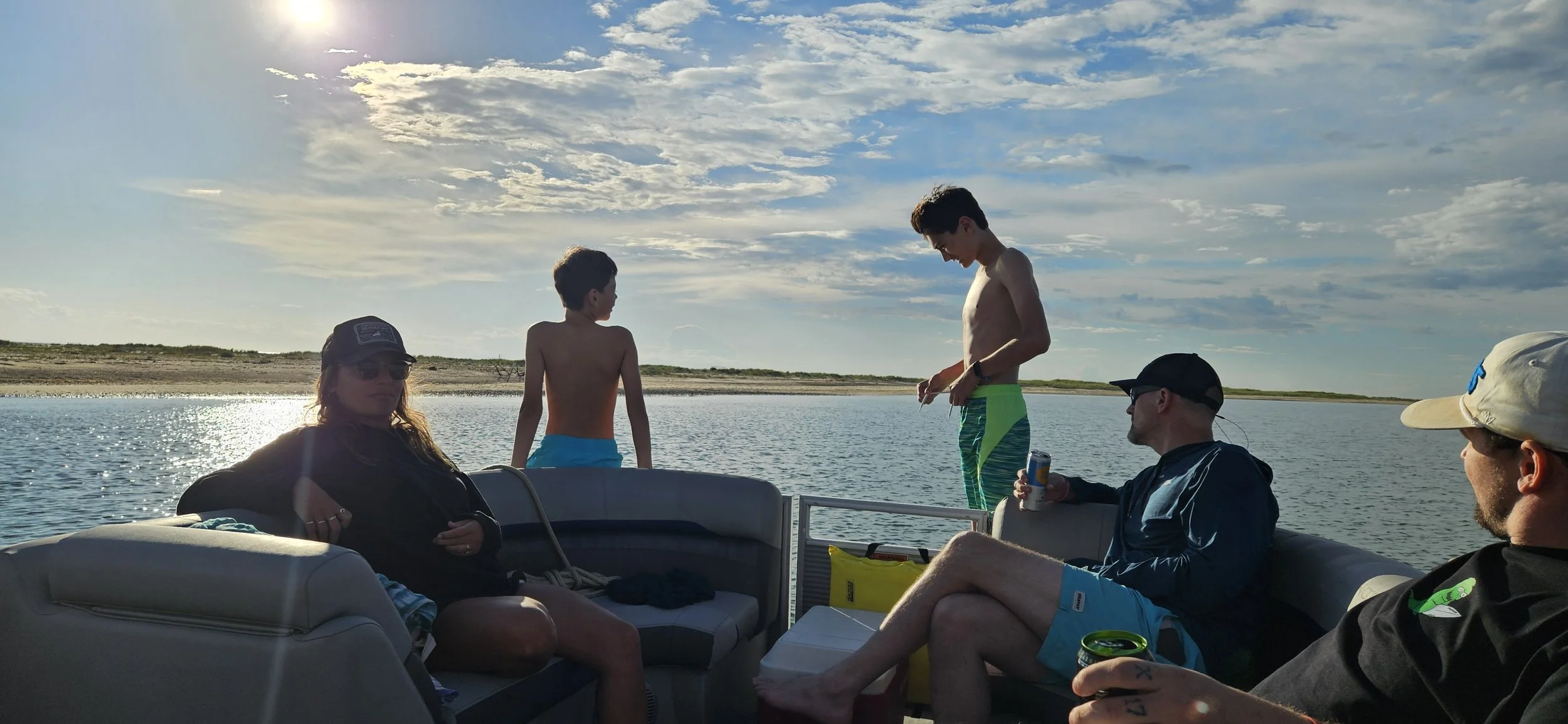 A group of five people on a boat enjoying a day on the water during sunset, with some sitting on the boat's seating and two standing by the edge, wearing swimwear and casual clothing.