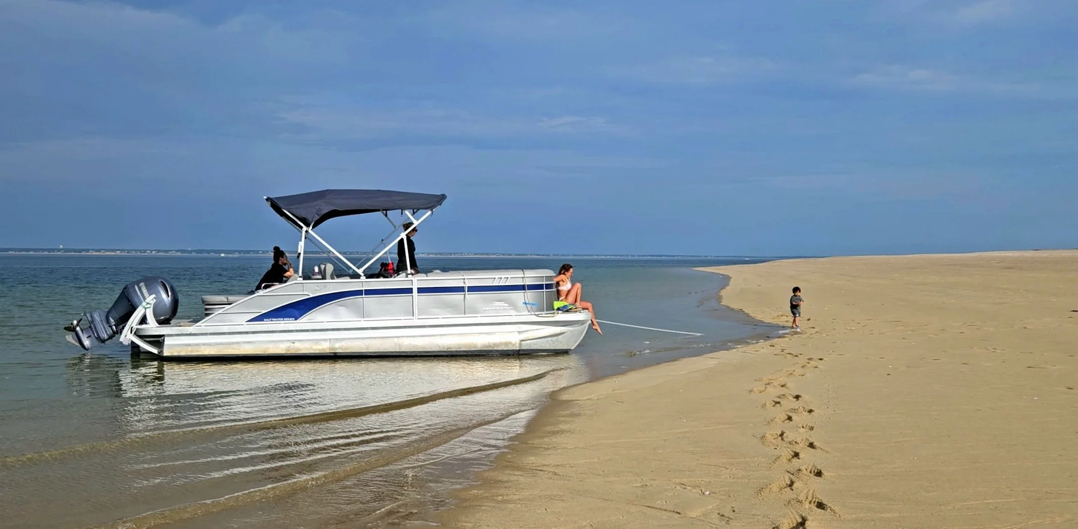 A boat anchored near the shore of a sandy beach with a woman sitting on the edge of the boat, a child playing nearby, and two people on the boat, under a cloudy sky.