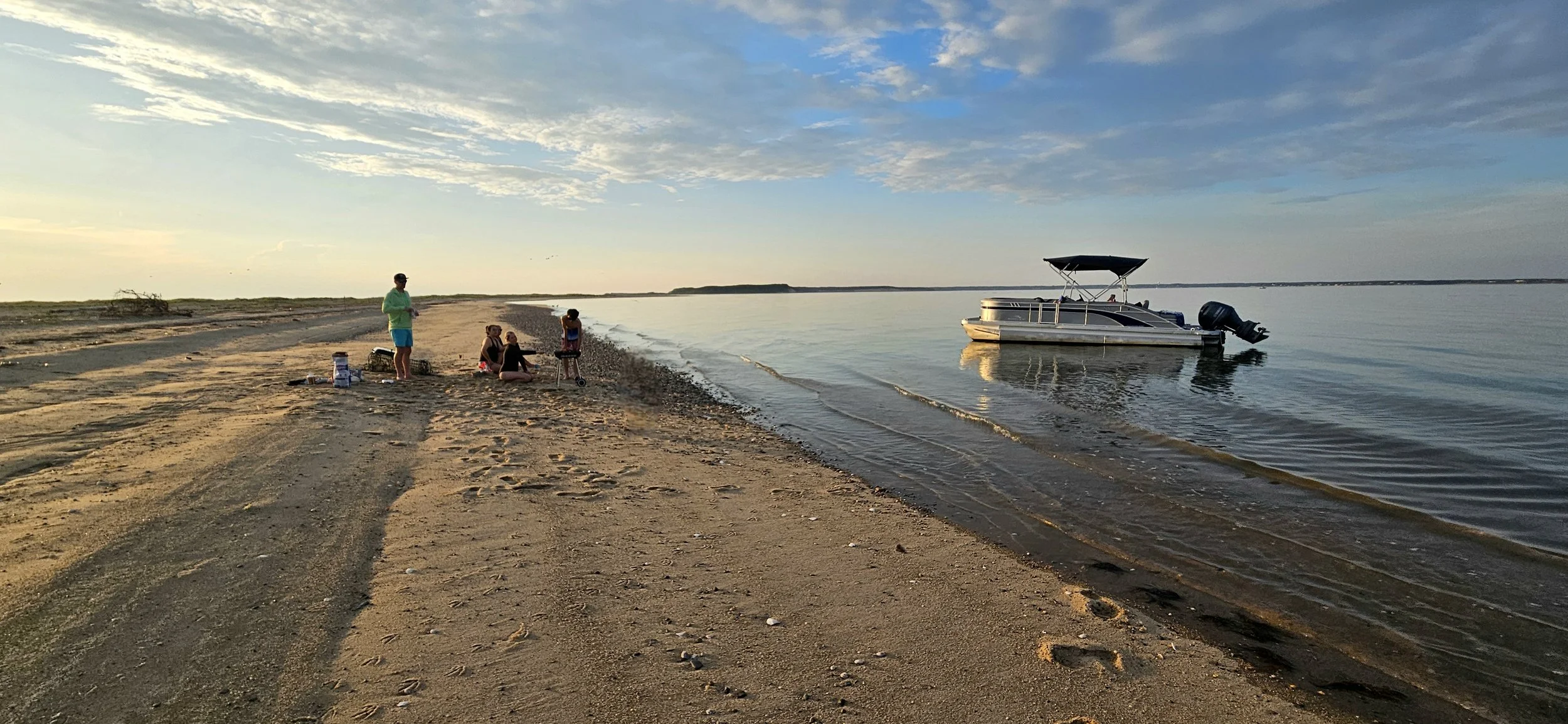 People relaxing on a sandy beach near a boat on calm water, under a partly cloudy sky during sunset.