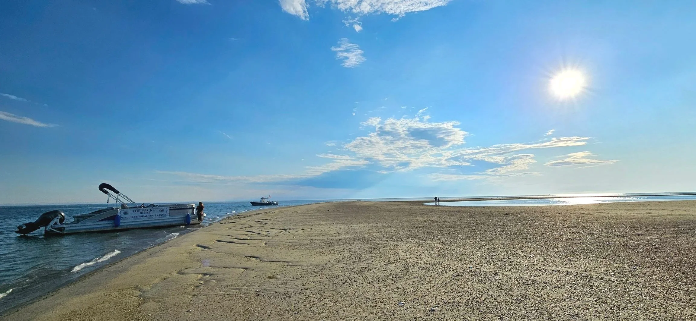 A sandy beach with a boat docked near the shore, a person standing beside it, and two people walking in the distance under a bright sun in a partly cloudy blue sky.