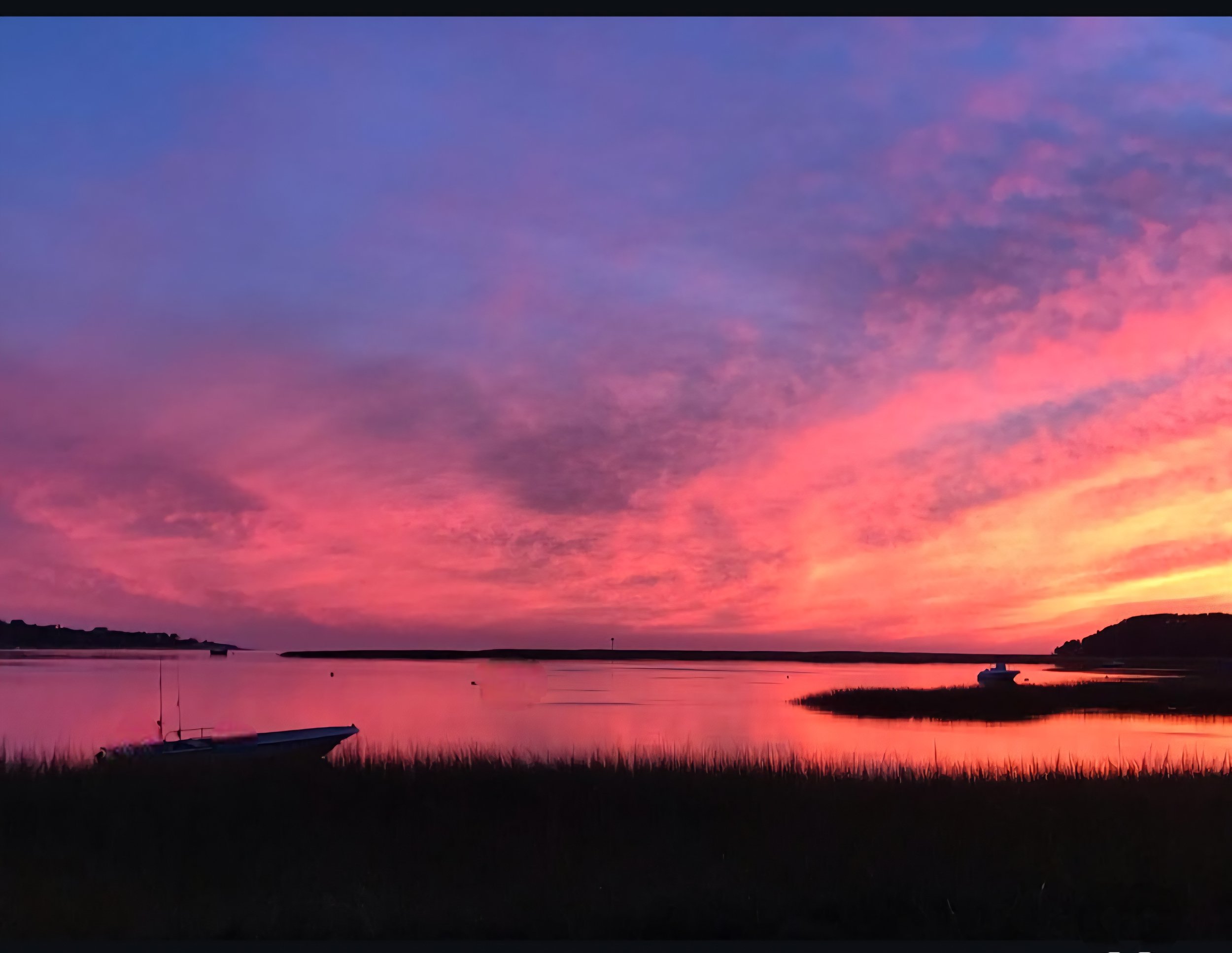 A vibrant sunset over a calm body of water with boats anchored near the shoreline, pink and purple clouds in the sky, and distant landforms on the horizon.
