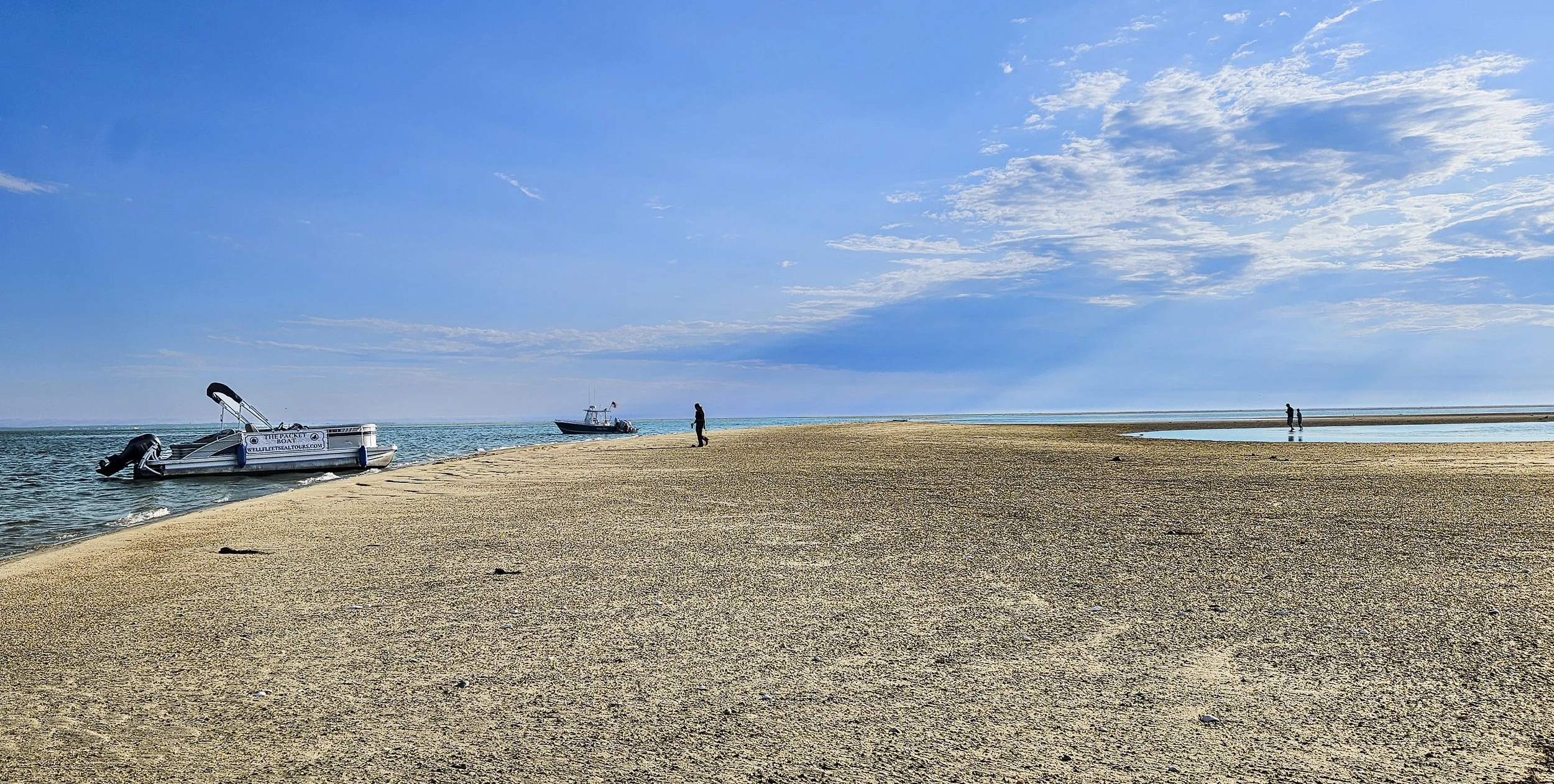A sandy beach with two boats, one anchored in the water and one heading out. A few people walk along the shoreline under a partly cloudy sky.