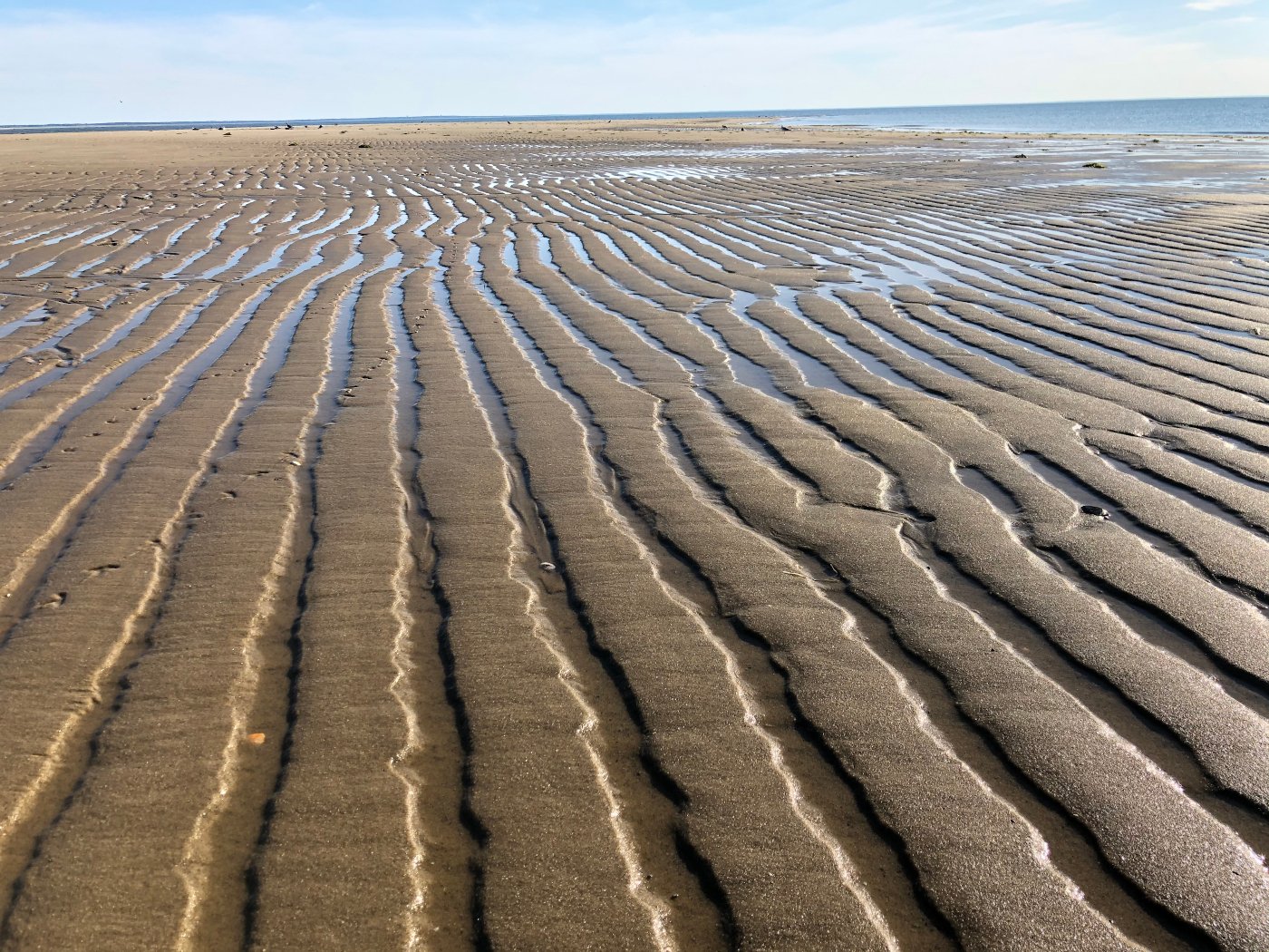 Sand with rhythmic ridges and shallow pools of water, extending towards the horizon at a beach, with the ocean in the background under a partly cloudy sky.