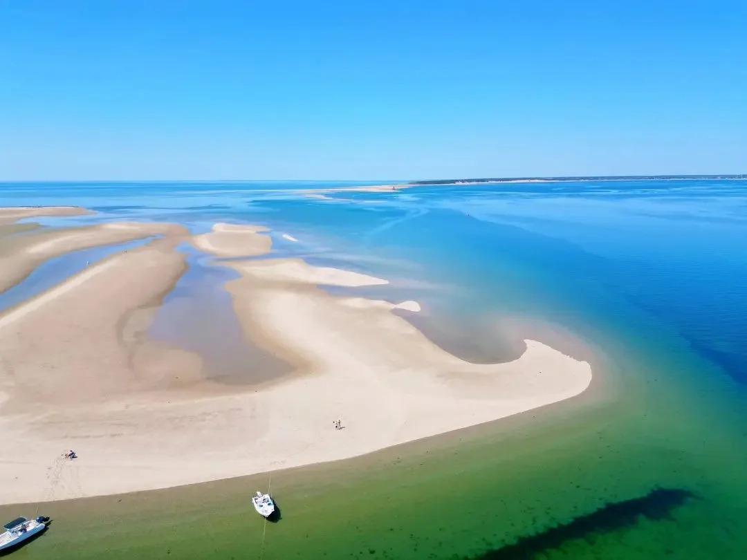 A beach with sandbars, boats, and clear blue water under a bright sky.