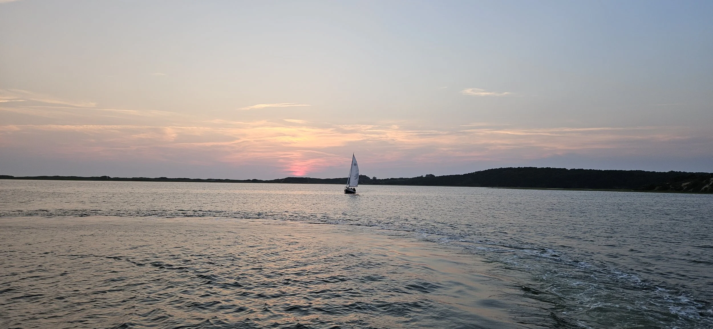 A sailboat on calm water during sunset with a partly cloudy sky and distant hills.