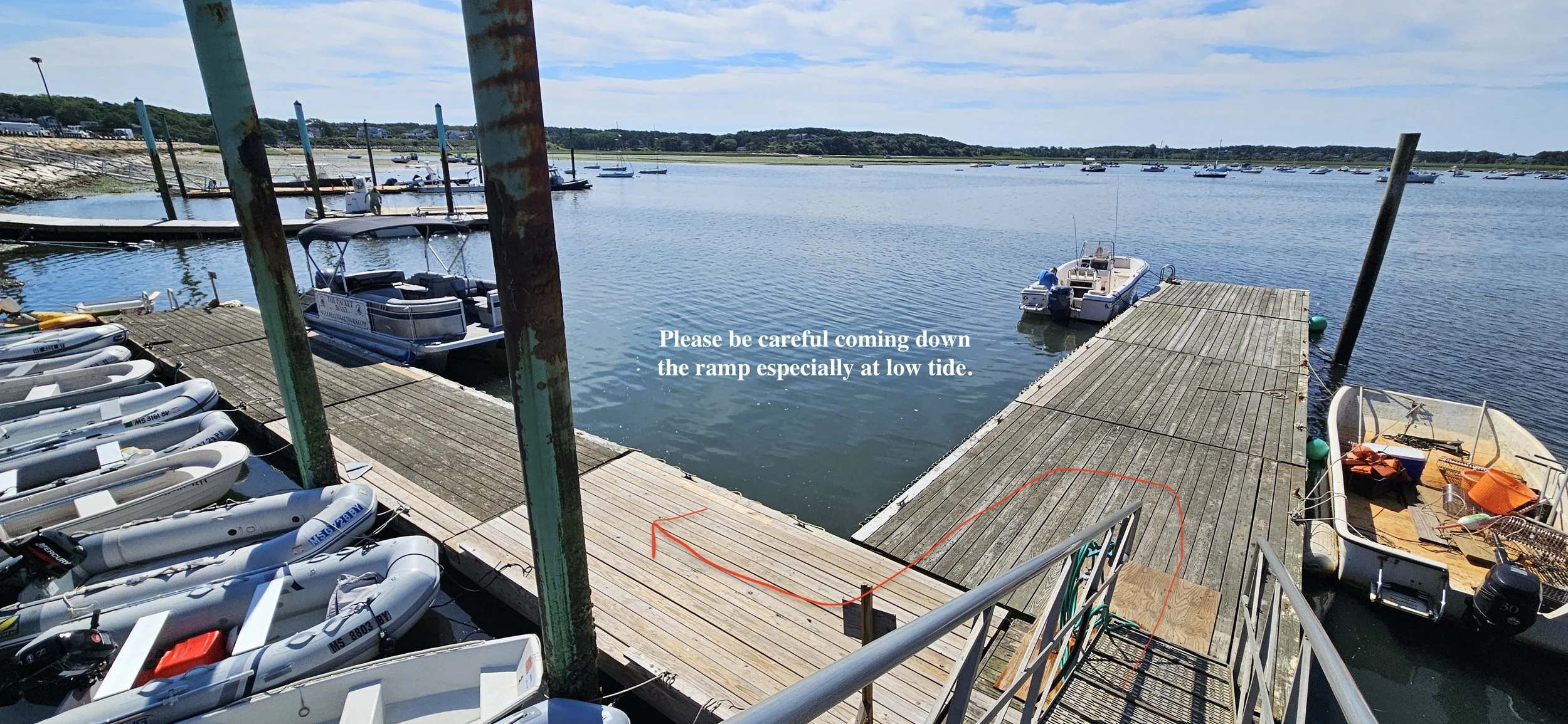 A marina with boats docked along a wooden ramp, some boats are inflatable. There is a warning sign that reads, 'Please be careful coming down the ramp especially at low tide.'