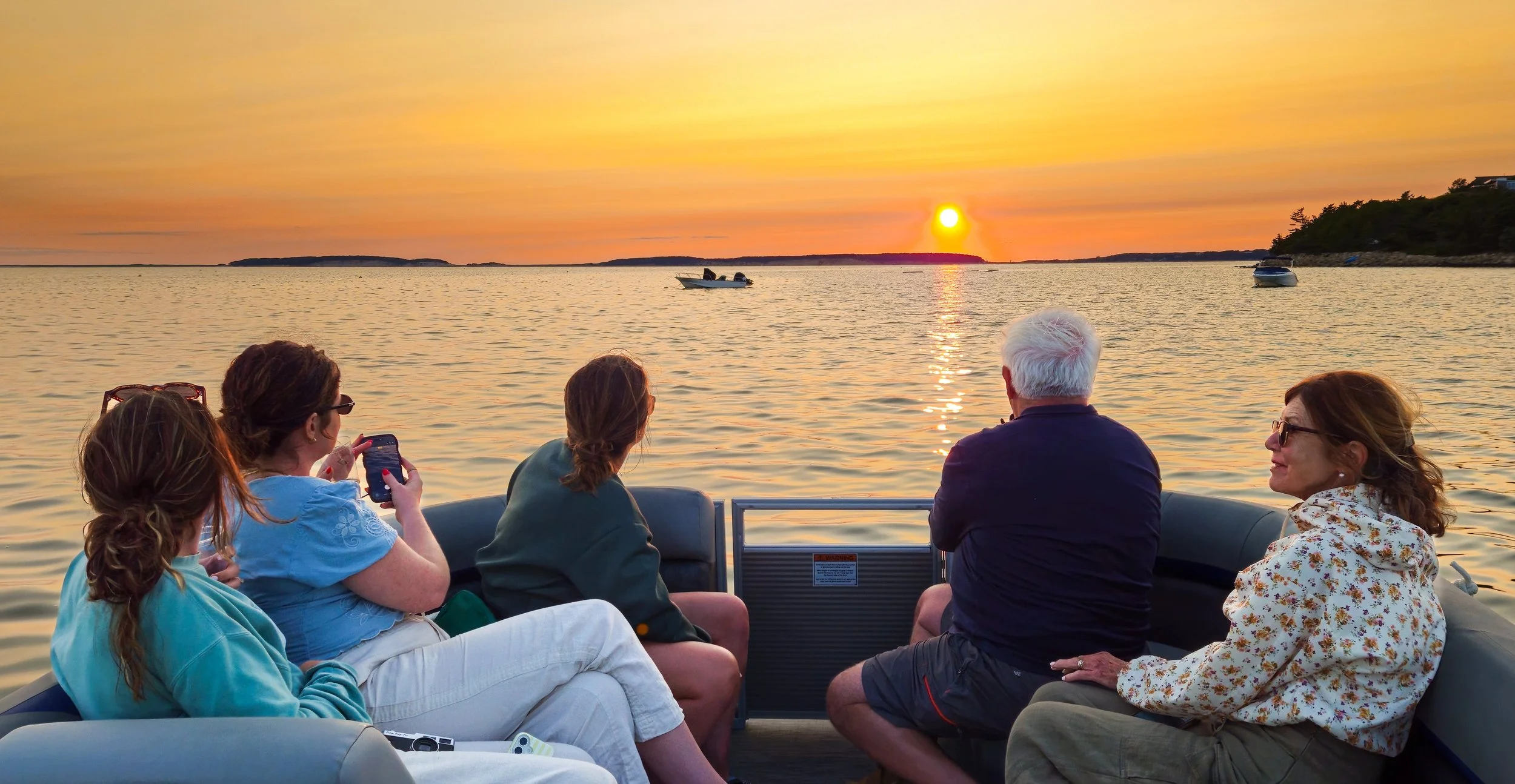 Group of five people enjoying a boat ride during sunset on a calm body of water, with boats and distant land on the horizon.