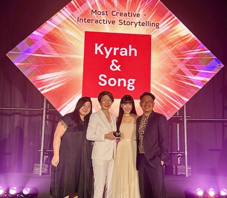 Four people standing on a stage in front of a large colorful display that reads 'Most Creative - Interactive Storytelling' and 'Kyrah & Song'. They are smiling, dressed in formal attire, and the woman second from the right is holding an award.