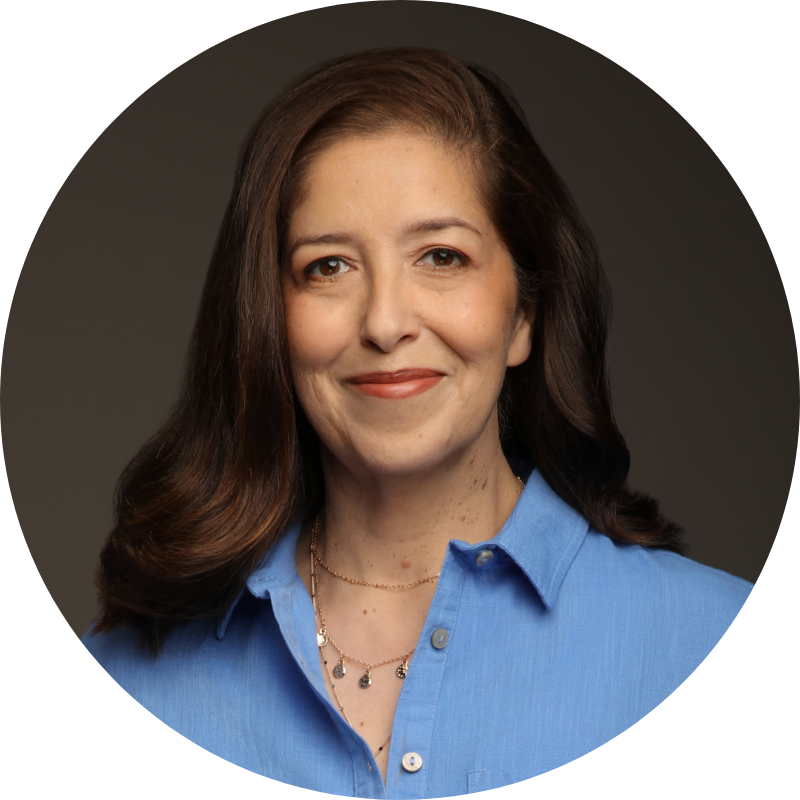 A woman with wavy brown hair smiling, wearing a blue shirt, against a dark background.