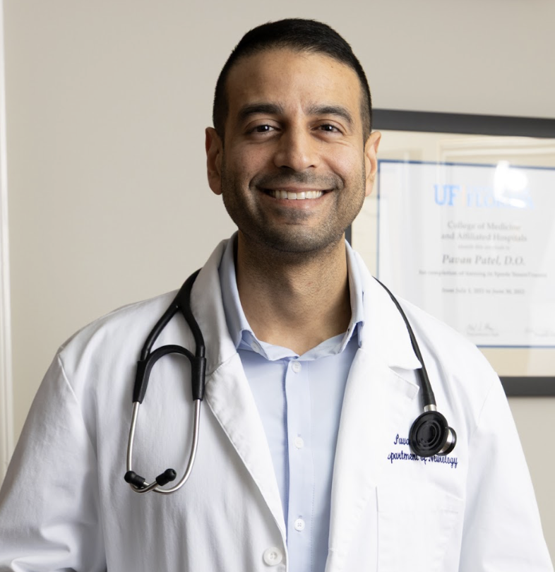 Smiling male doctor in white coat with stethoscope around neck, standing in medical office with framed certificate in background.