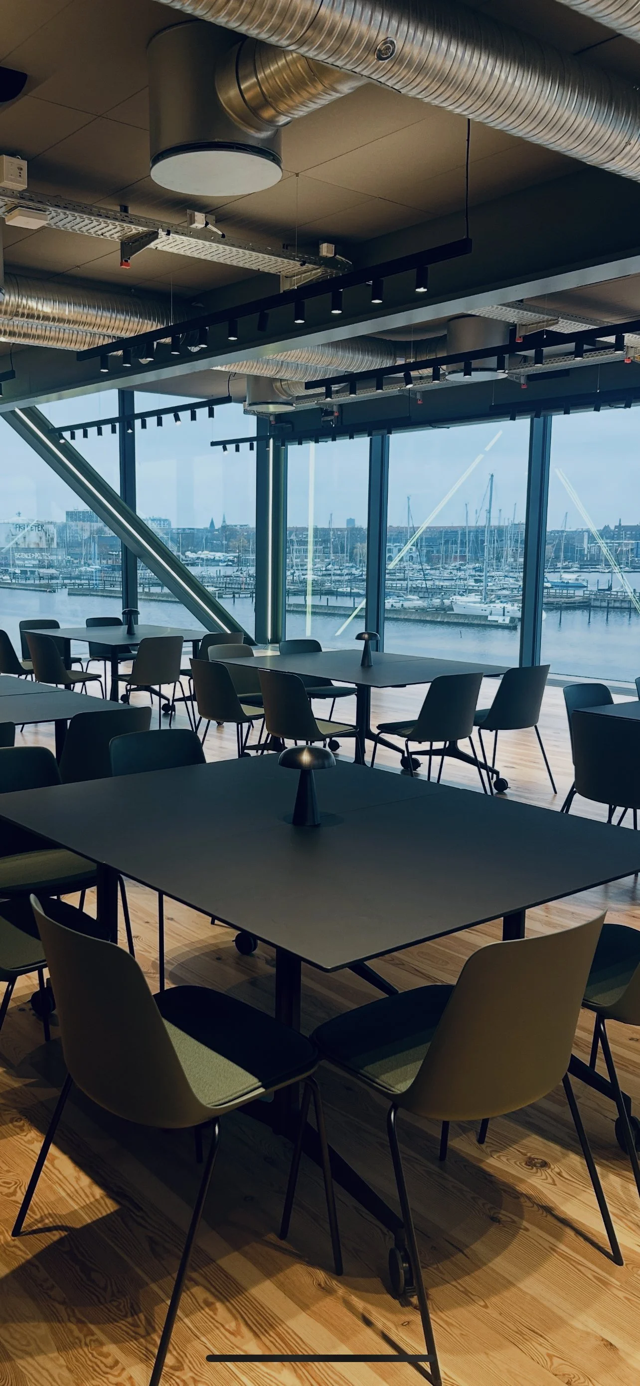 Modern indoor dining area with several tables and chairs near large windows overlooking a marina with sailboats, exposed industrial ceiling ducts, and track lighting. Holm8