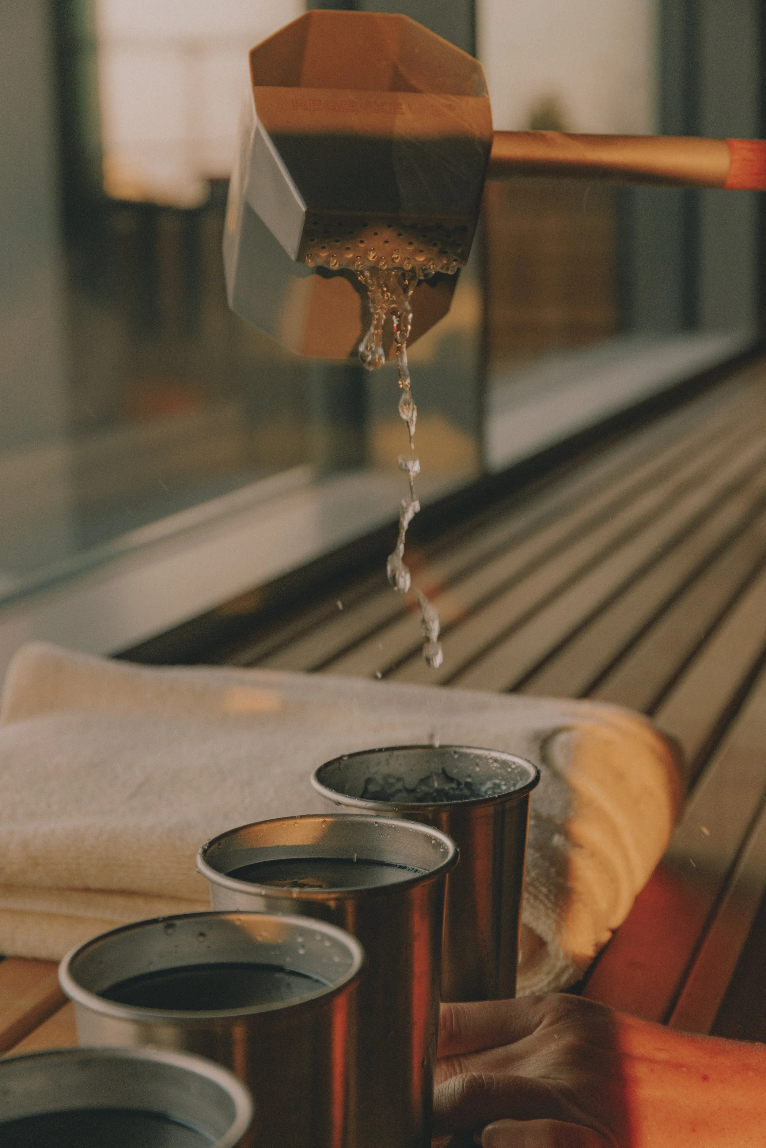 A saunagus pot is pouring hot water into metal cups on a wooden table, with a beige towel and a glass window in the background. Holm8