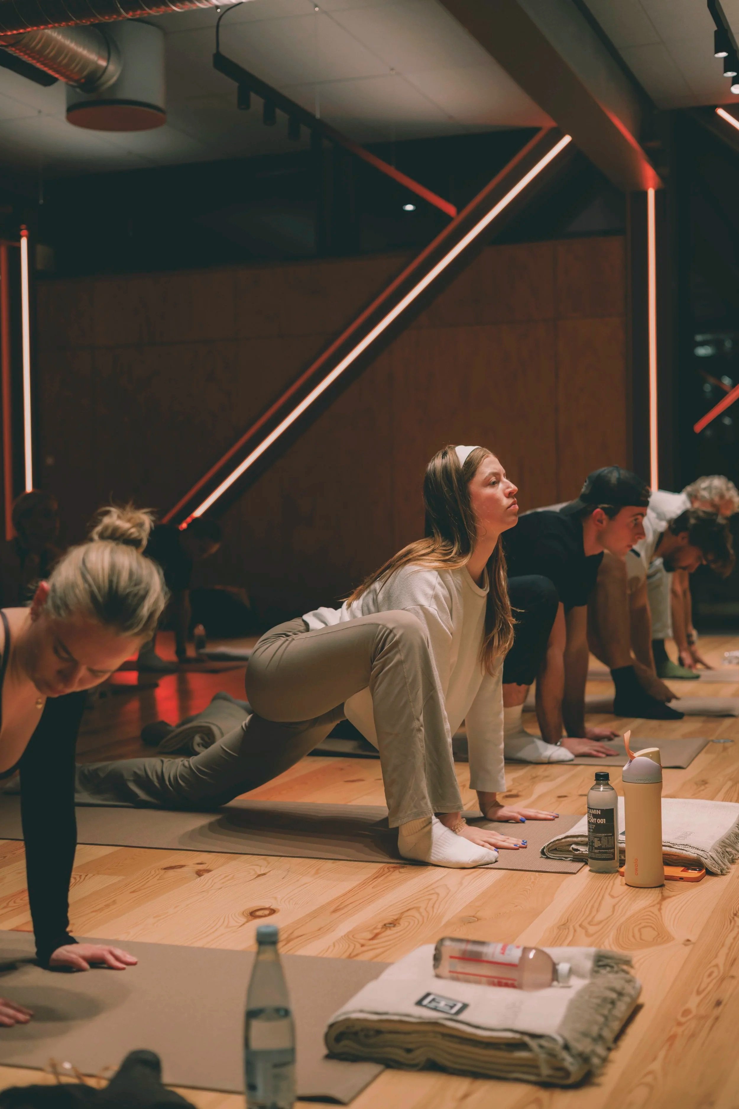 People participating in a yoga class, performing a plank pose on yoga mats in a dimly lit studio with contemporary lighting and wooden floors. Holm8