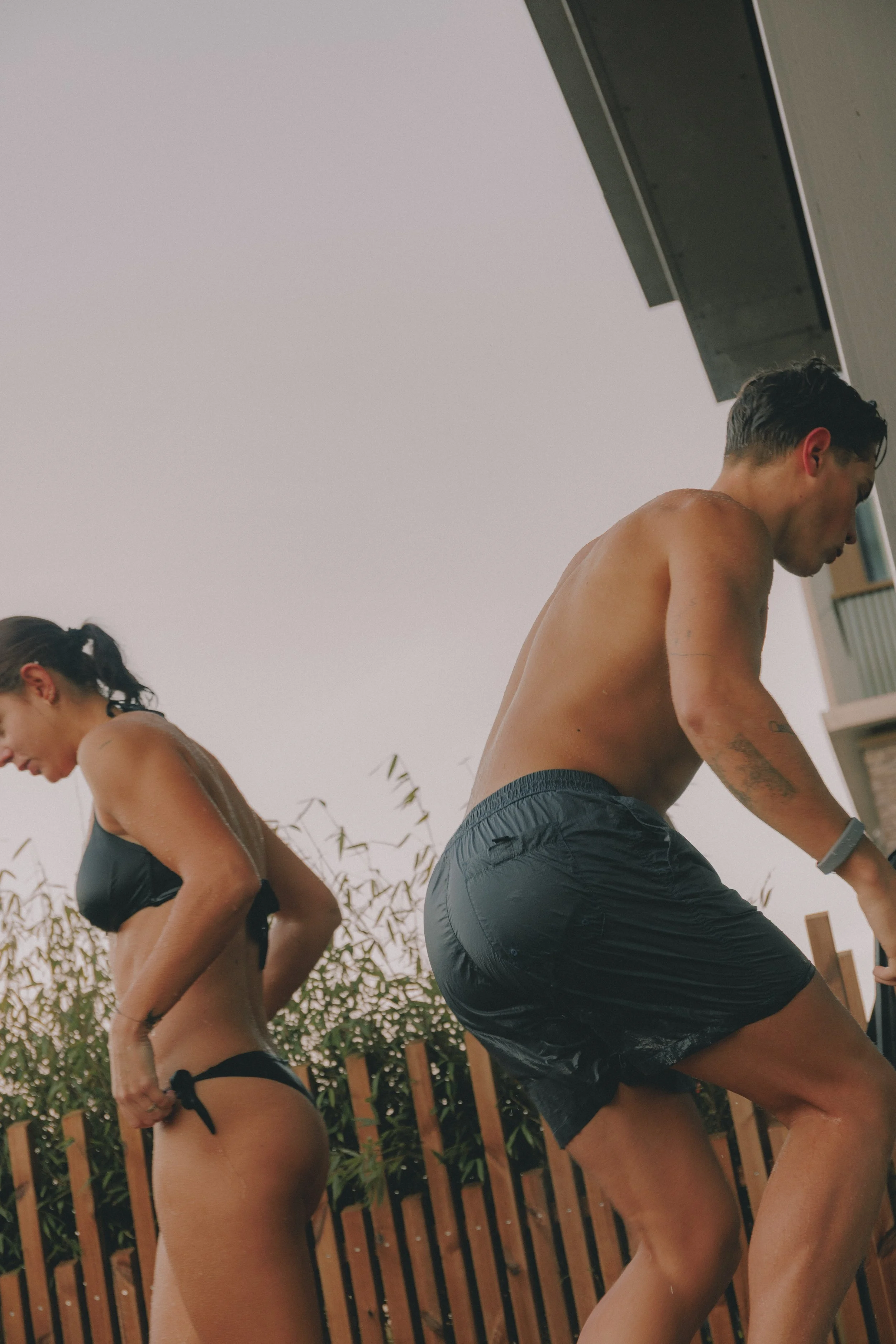 A man and a woman in swimwear pouring water on each other outdoors near a wooden fence, with plants and a building in the background. Holm8