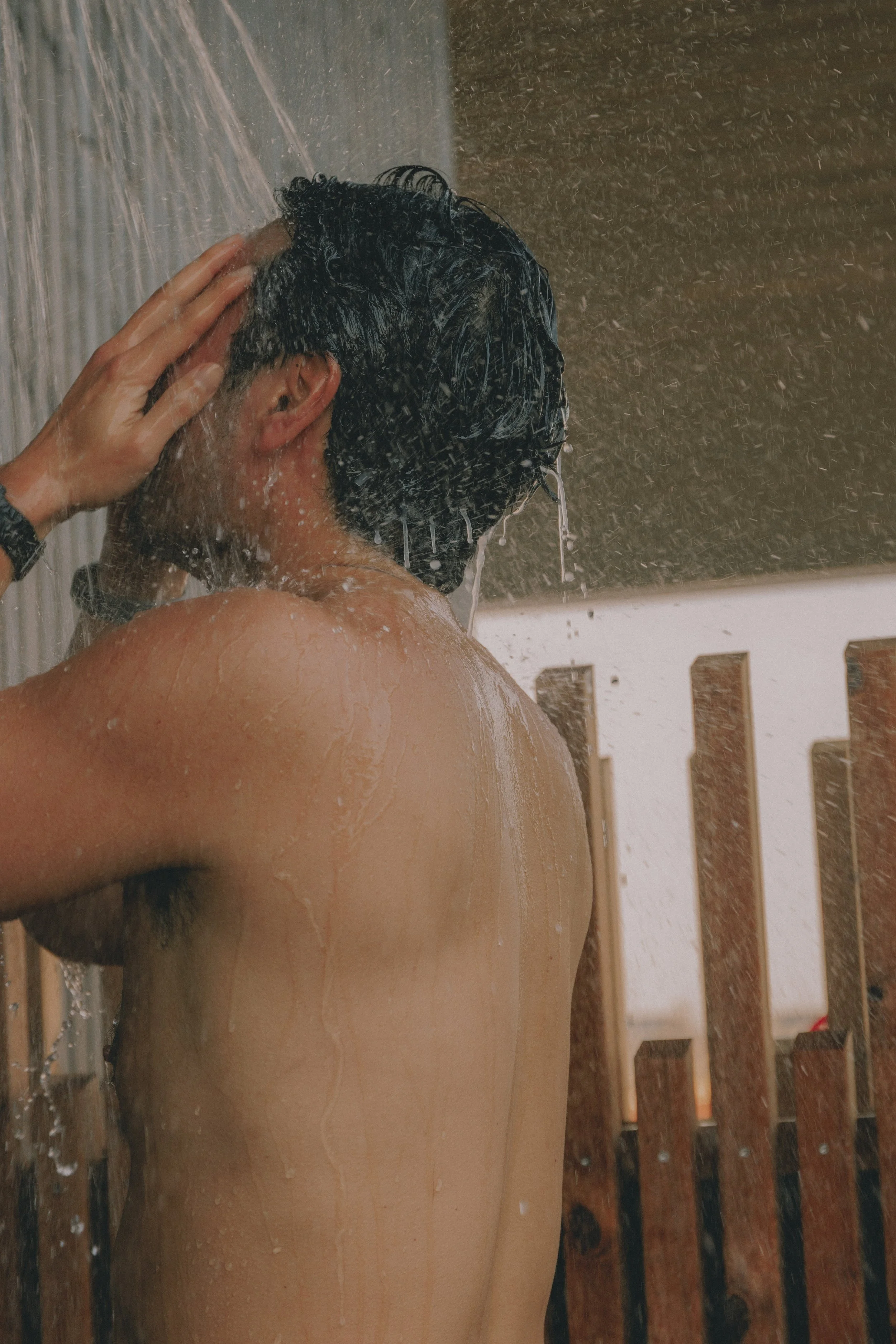 A man with dark hair taking a shower, with water splashing and running down his back. Holm8