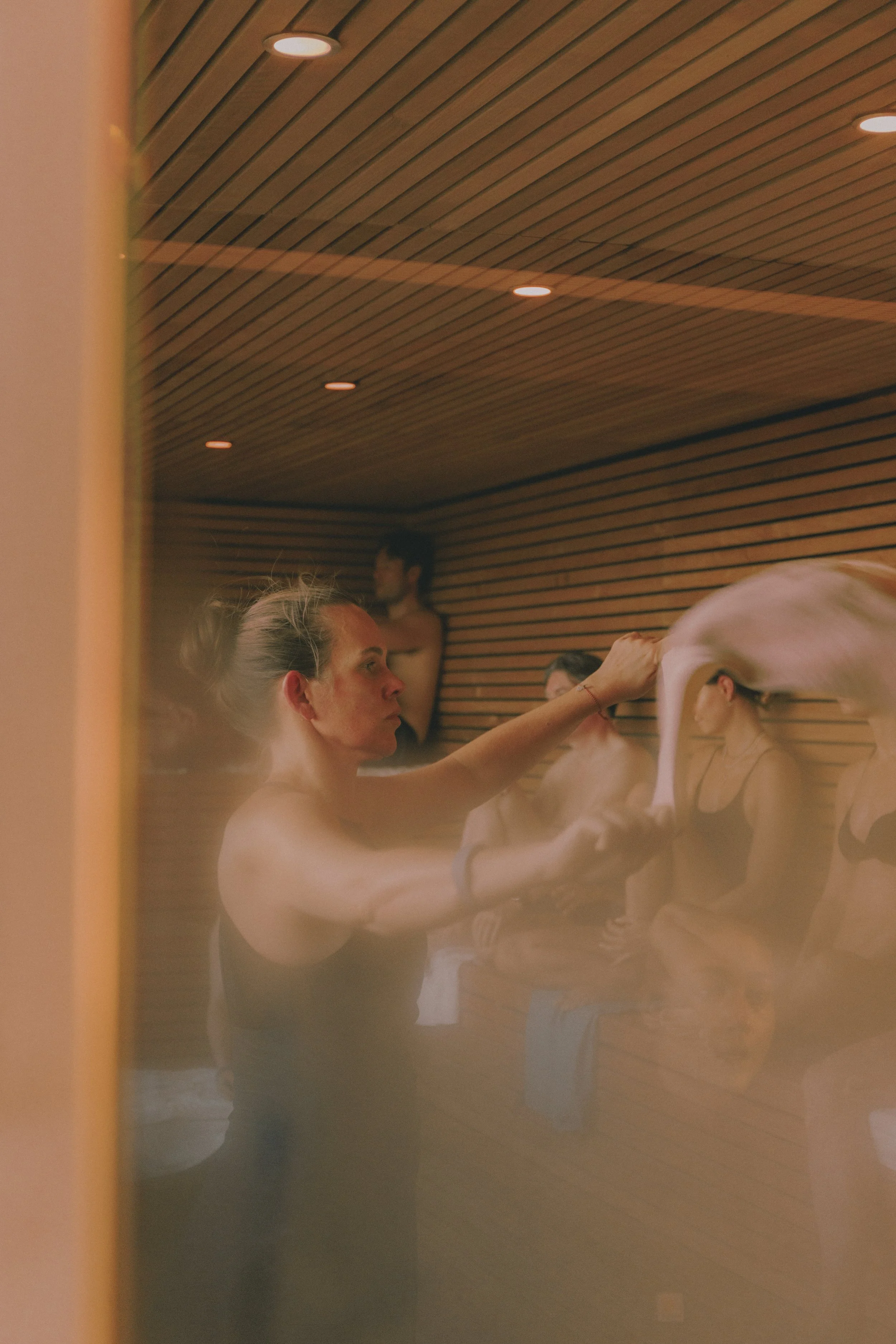 A woman in a sauna is tending to other women, who are stretching and adjusting their hair, with a wooden interior and warm lighting. Holm8