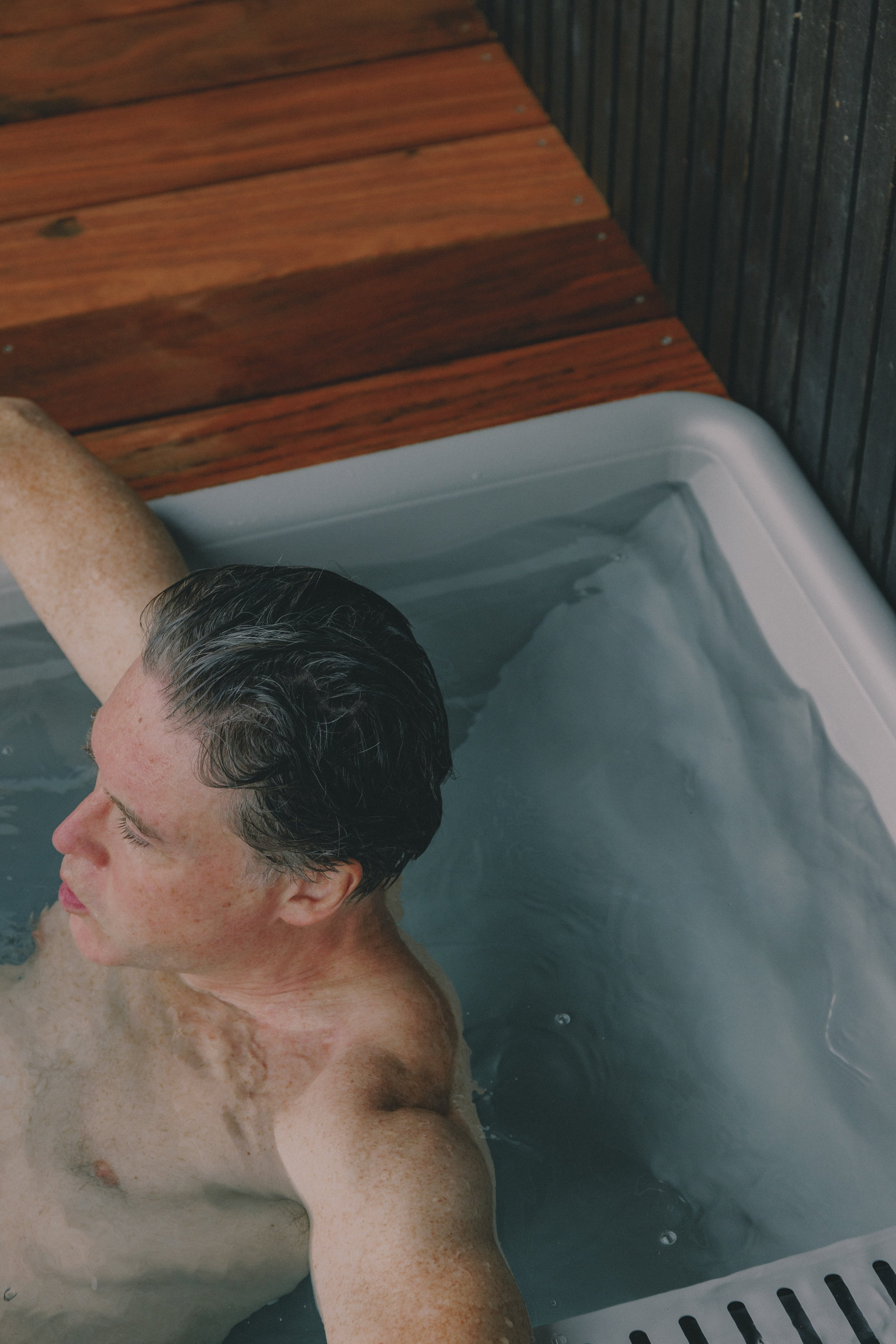 A man with dark, slicked-back hair relaxing in an outdoor cold plunge with water at his chest, surrounded by wooden decking and a dark wooden fence. Holm8