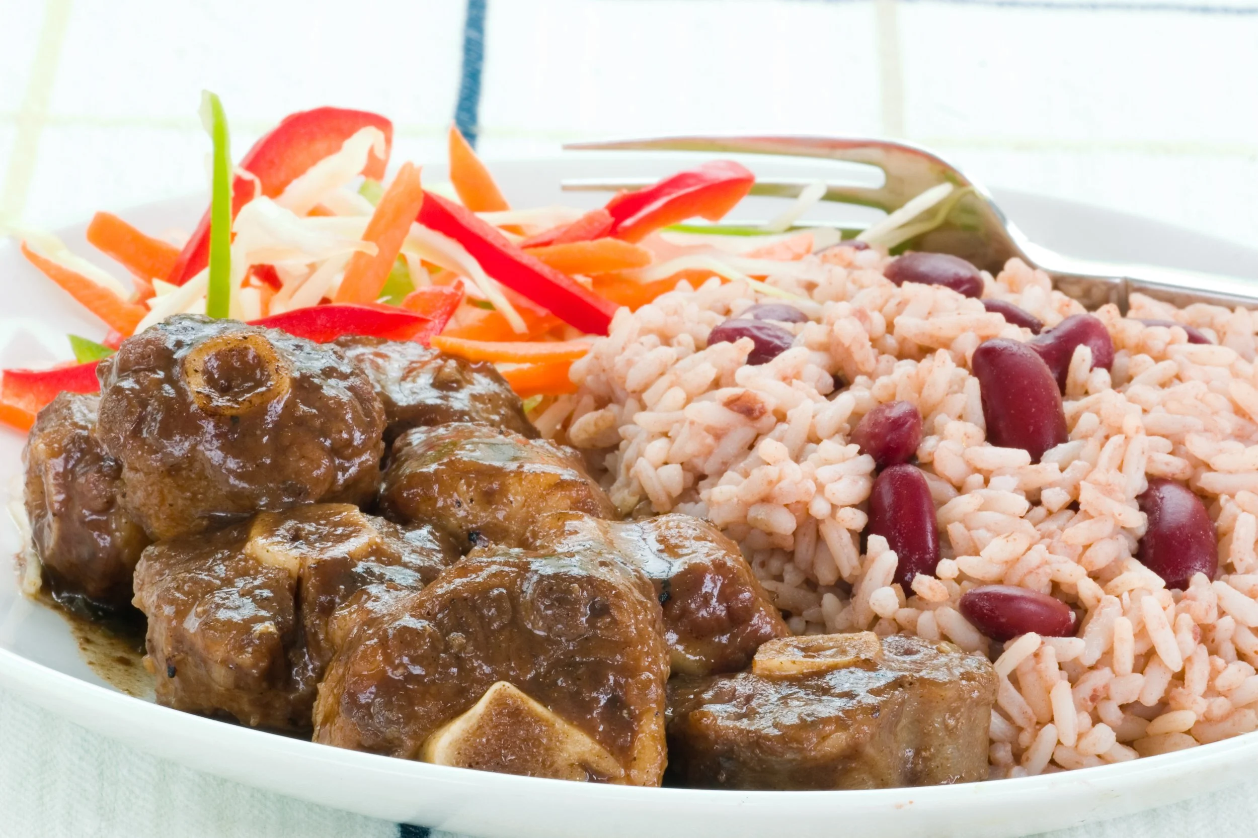 Plate of oxtail with rice and vegetables, including shredded carrots, sliced red bell peppers, shredded cabbage, and kidney beans.