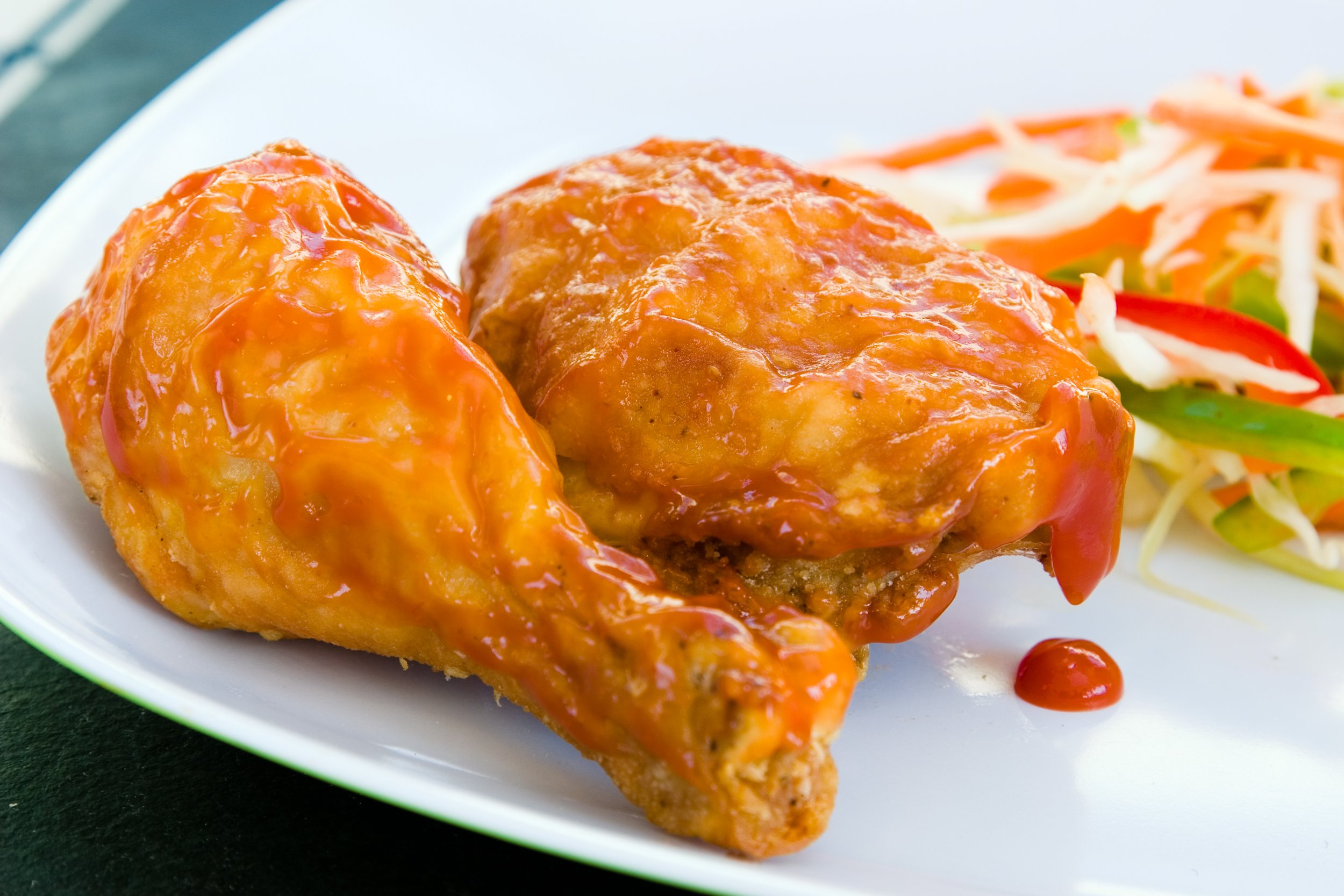 Two pieces of fried chicken drumsticks covered in fried chicken (red, savoury) sauce, served with a side of coleslaw salad containing shredded cabbage, carrots, and sliced red and green bell peppers on a white plate.