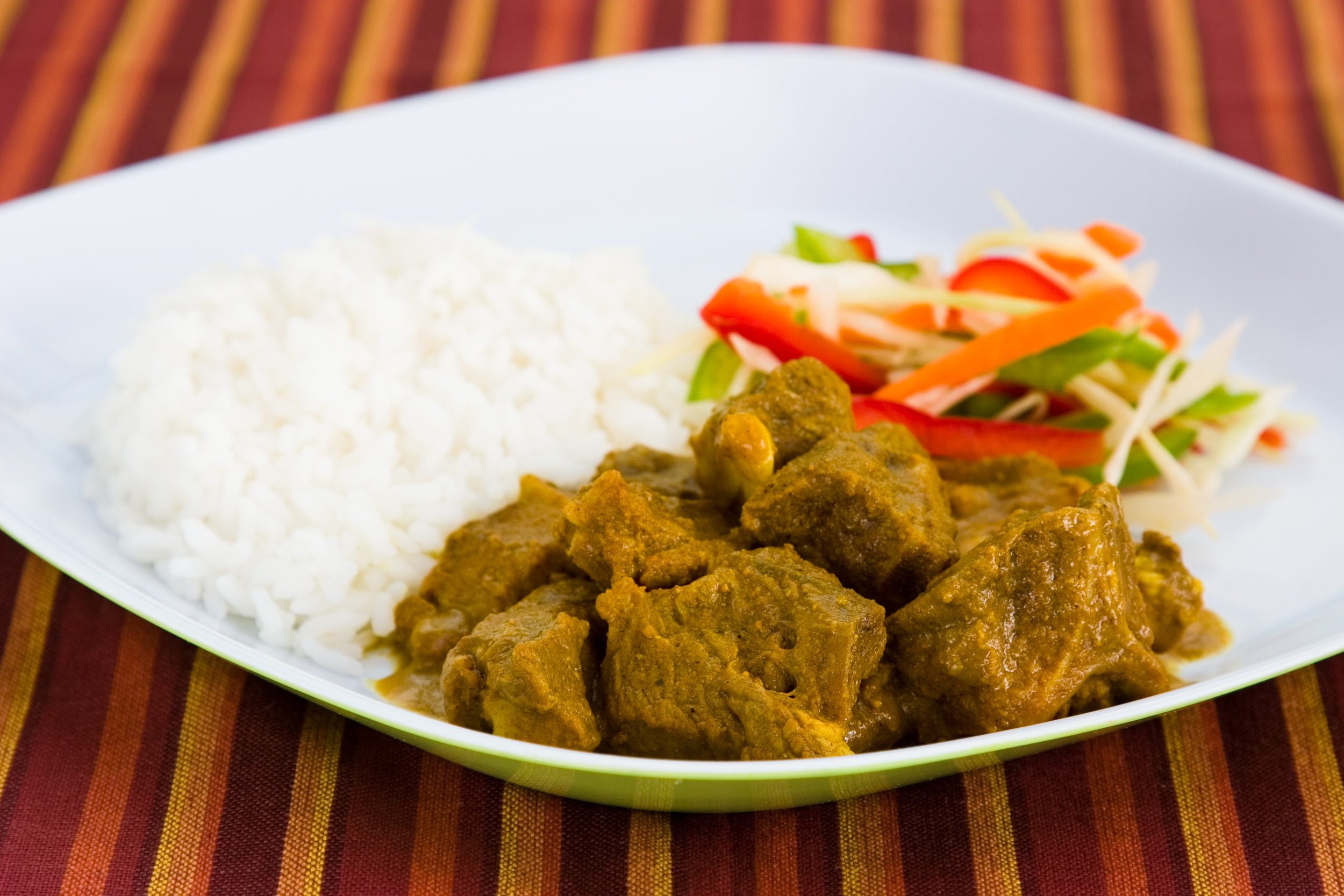 Plate with white rice, curry goat, and a side of colourful coleslaw on a striped tablecloth.