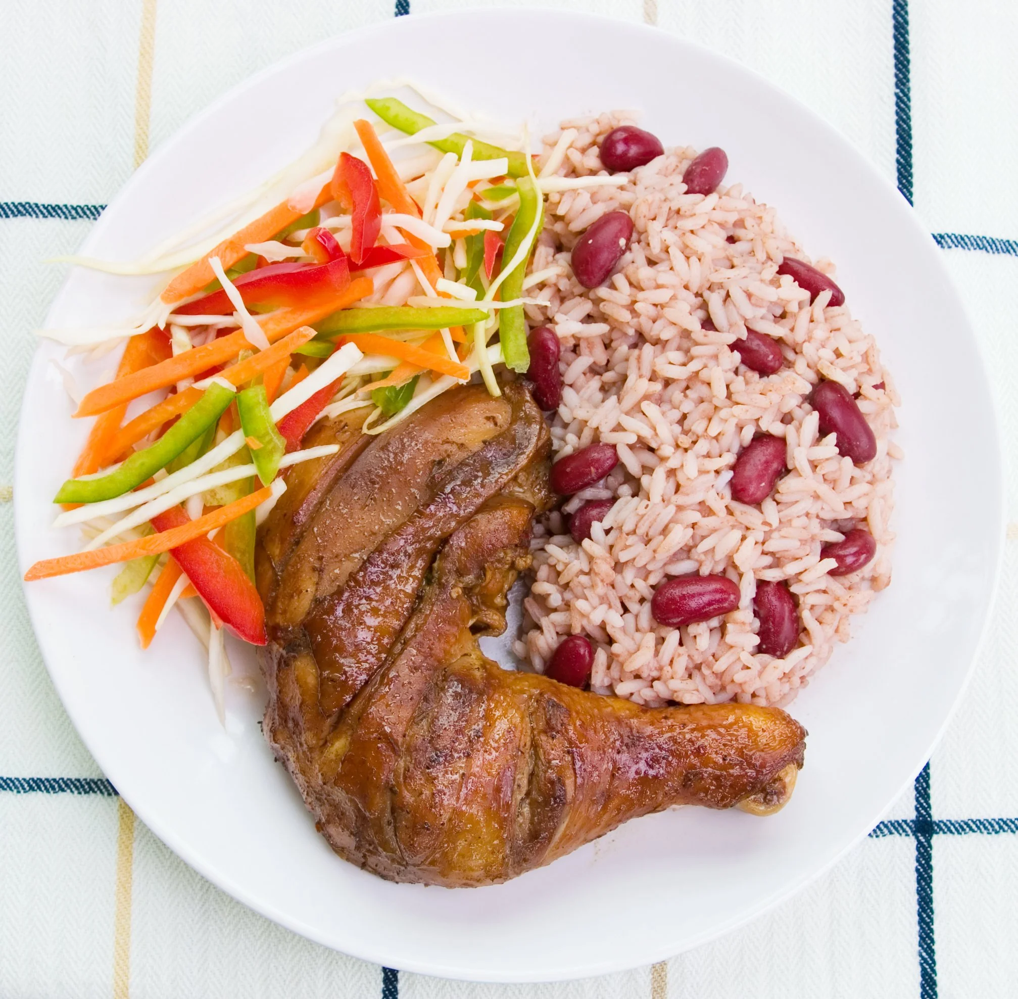 Plate with grilled jerk chicken leg, rice and peas (kidney beans), coleslaw with carrots and green pepper slices.