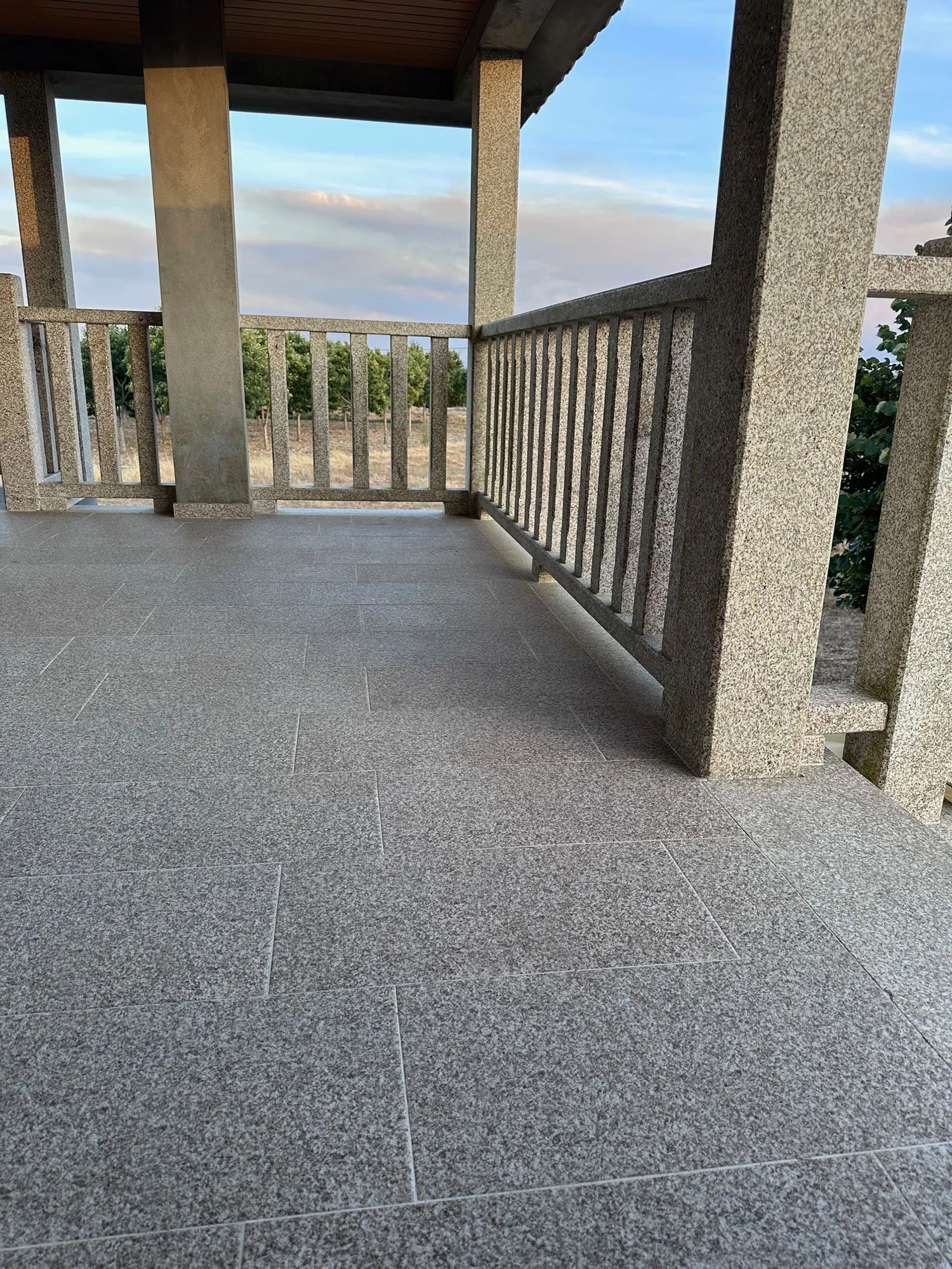 Balcon avec sol en carreaux gris, murs en pierre grise, vue sur un paysage rural avec des arbres et un ciel partiellement nuageux.