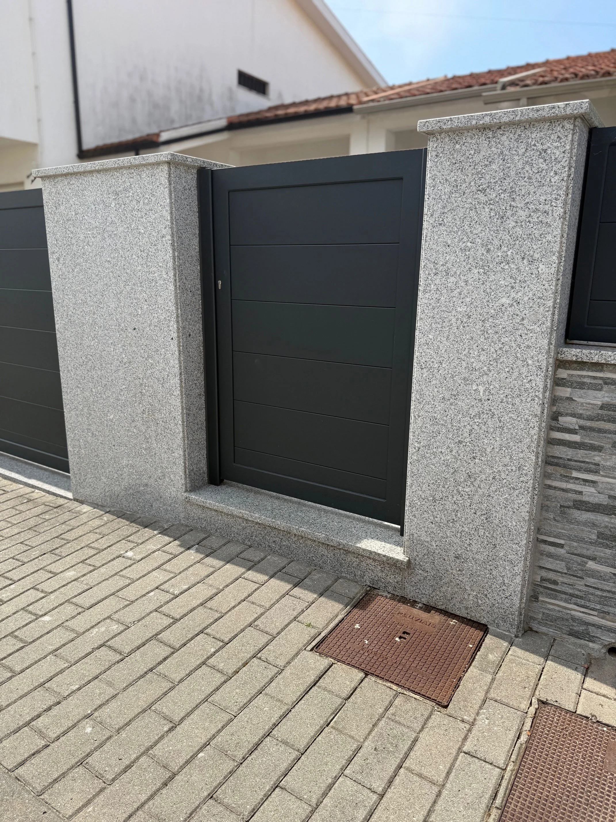 Une clôture en métal noir avec un portail dans un mur en pierre gris clair, pavé en béton en bas, devant une maison avec un toit en tuiles rouges.