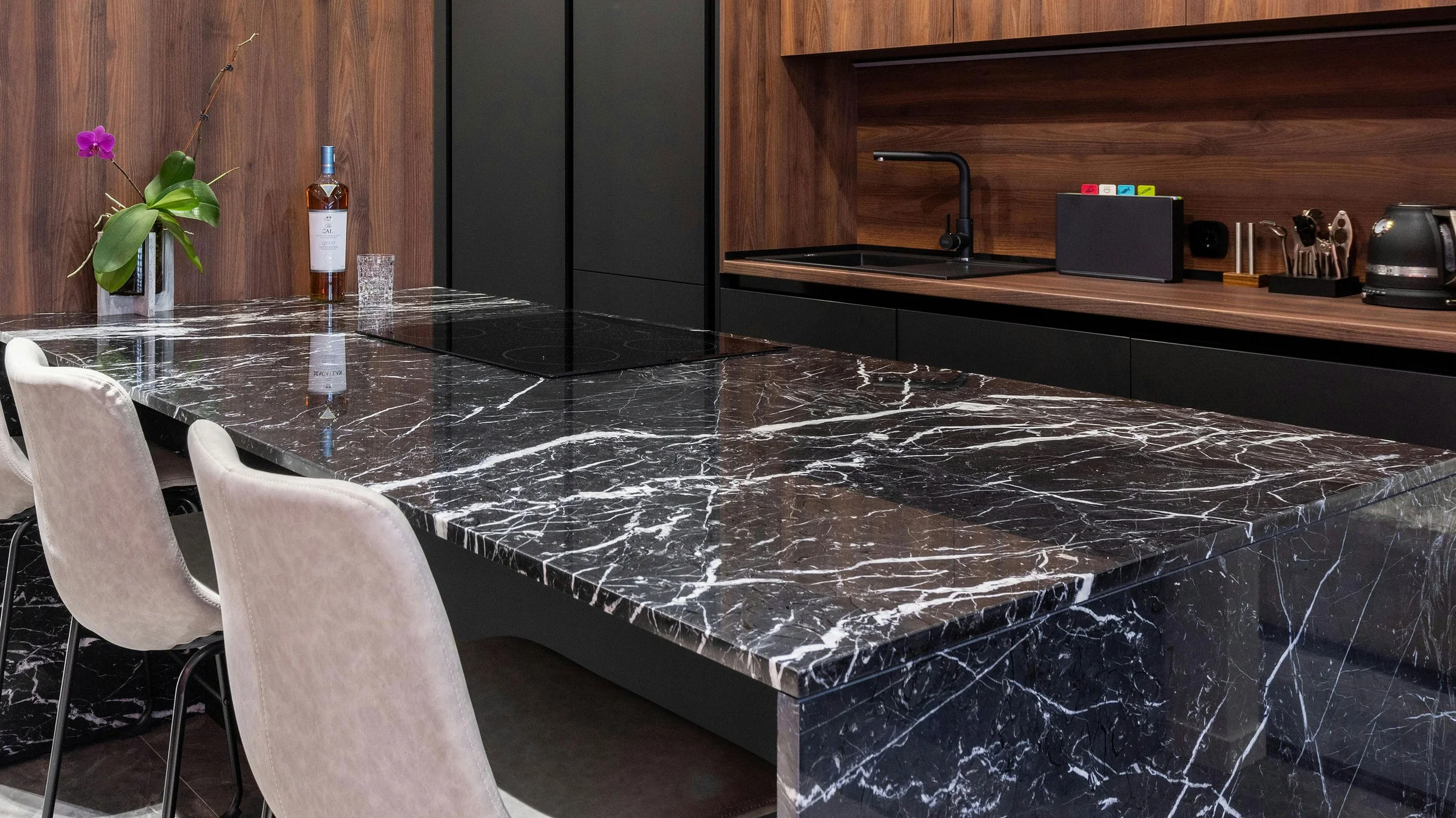 Cuisine moderne avec plan de travail en marbre noir avec veines blanches, comptoir en bois, évier noir, et appareils électroménagers noirs et en acier en arrière-plan, avec des chaises en tissu clair autour du plan de travail.