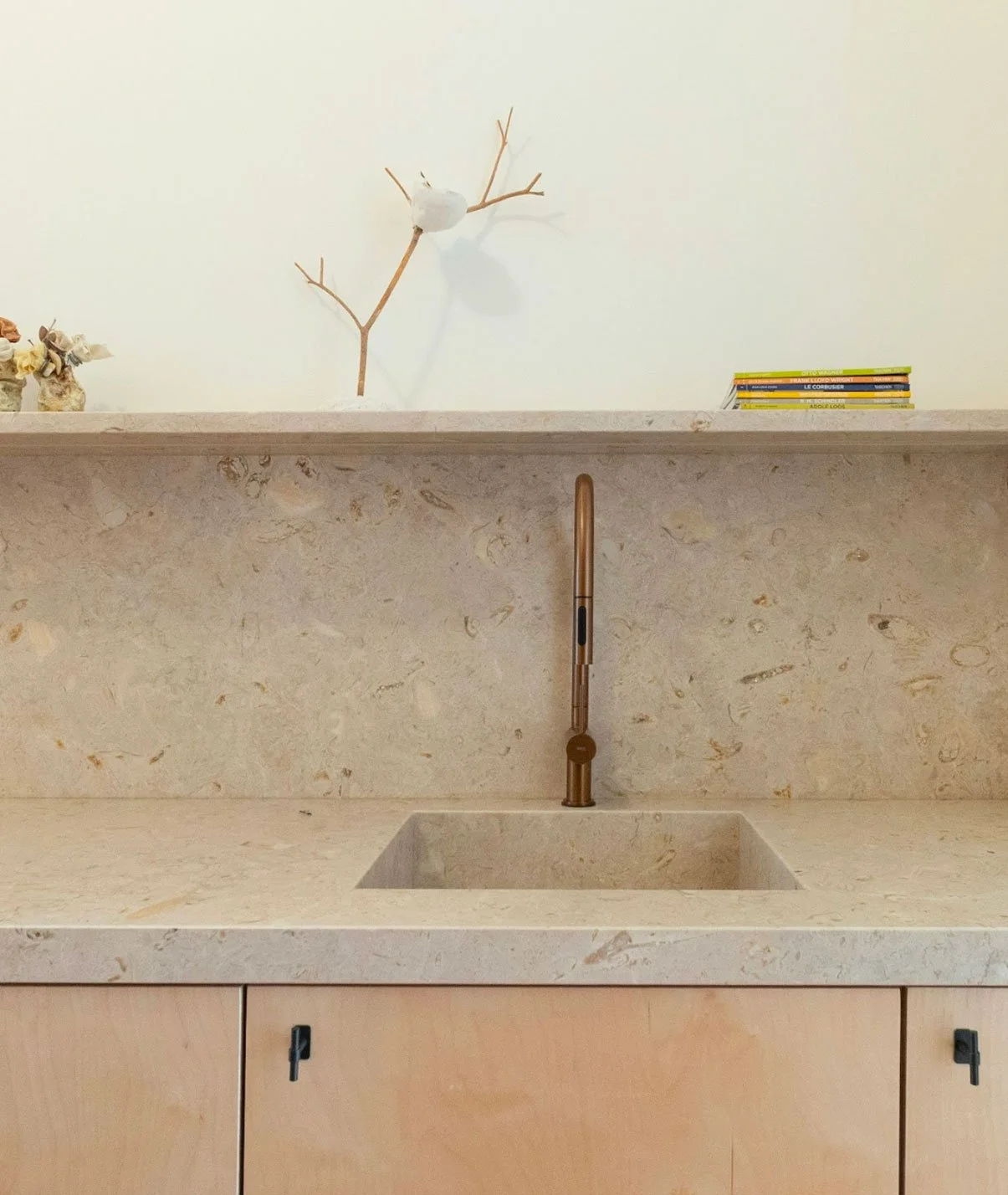 Plan de travail en pierre avec évier intégré et robinet en cuivre, étagère en pierre au-dessus avec des livres et décorations, mur blanc au fond.