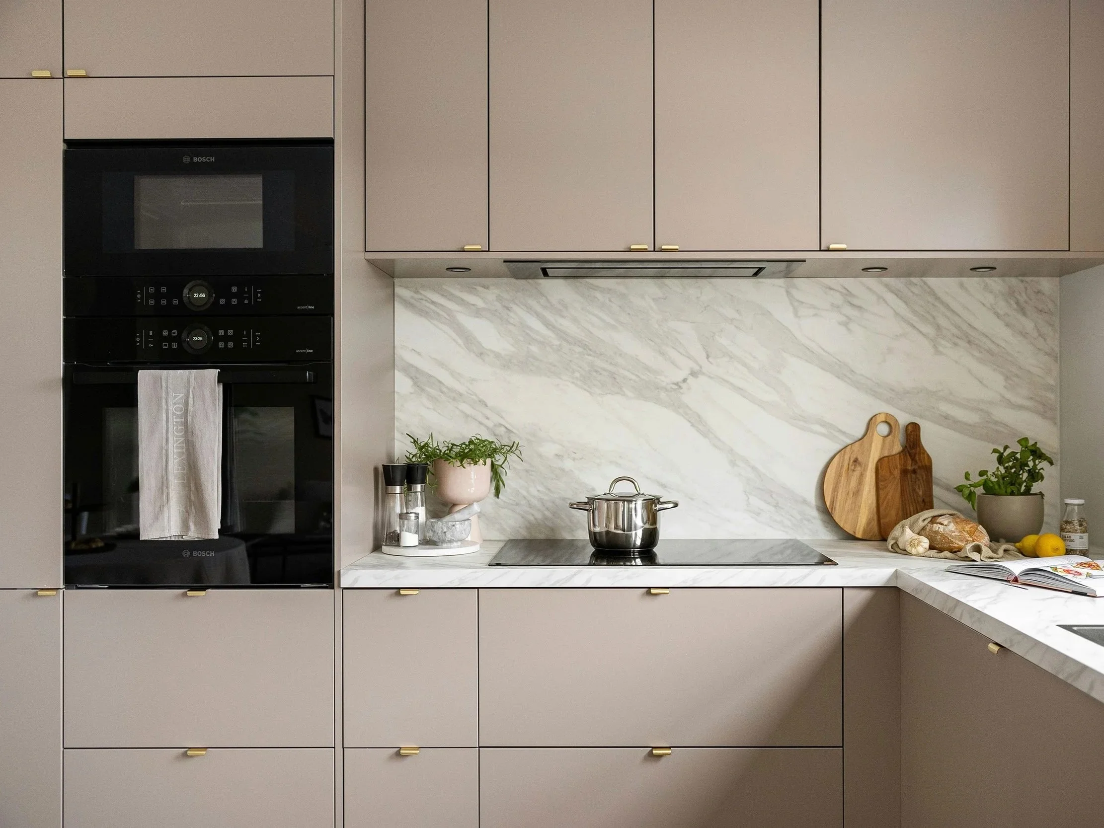 Cuisine moderne avec comptoir en marbre, placards beige, et mur en marbre, avec presse-purée, plantes, planche à découper, pain, livre de cuisine, et citron.