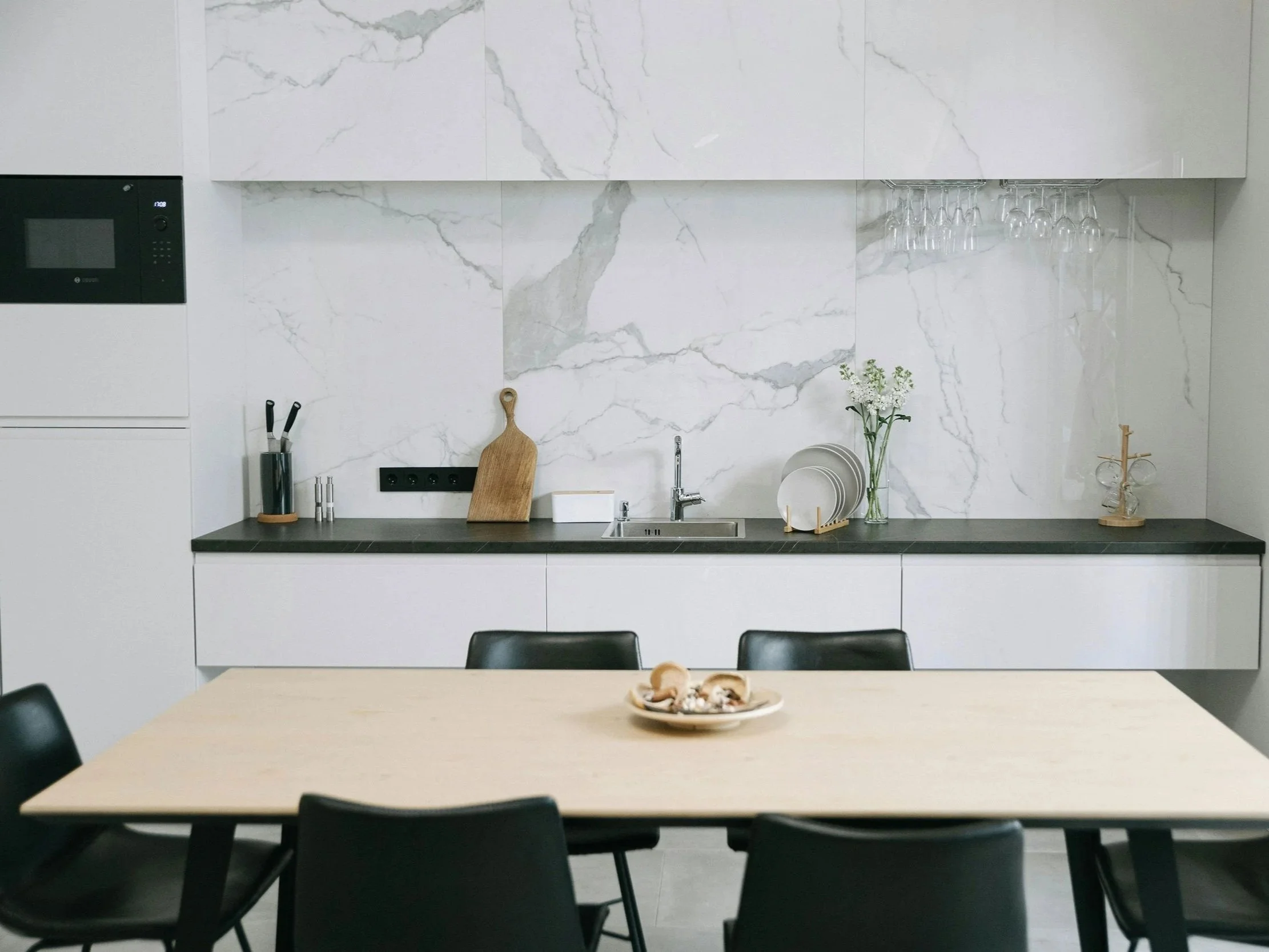 Cuisine moderne avec un plan de travail noir, une évier, des assiettes blanches, un vase avec des fleurs, une tablette à verres, et des décorations en bois, avec une table en bois clair entourée de chaises noires