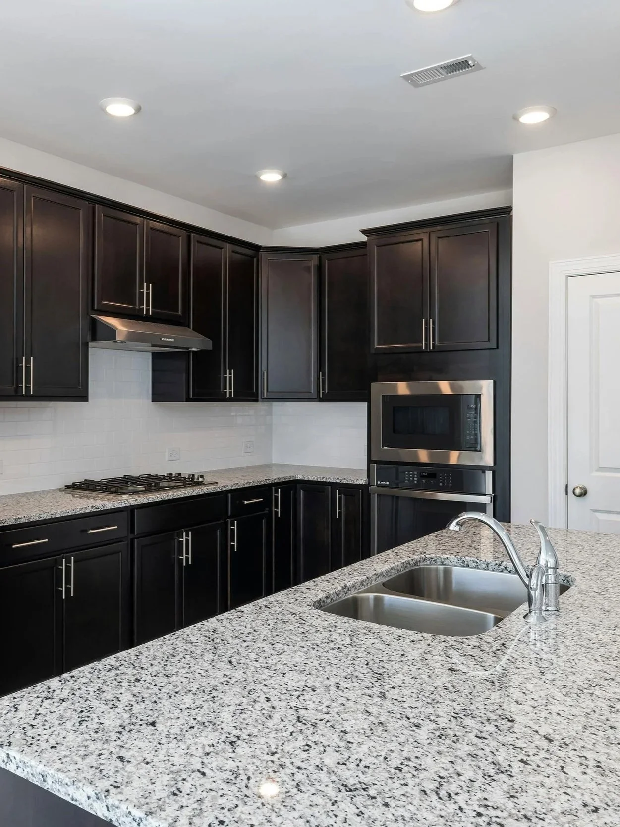 Cuisine moderne avec plans de travail en granite, armoires en bois foncé, évier double en acier inoxydable, four micro-ondes intégré, et cuisinière à gaz, éclairage encastré.