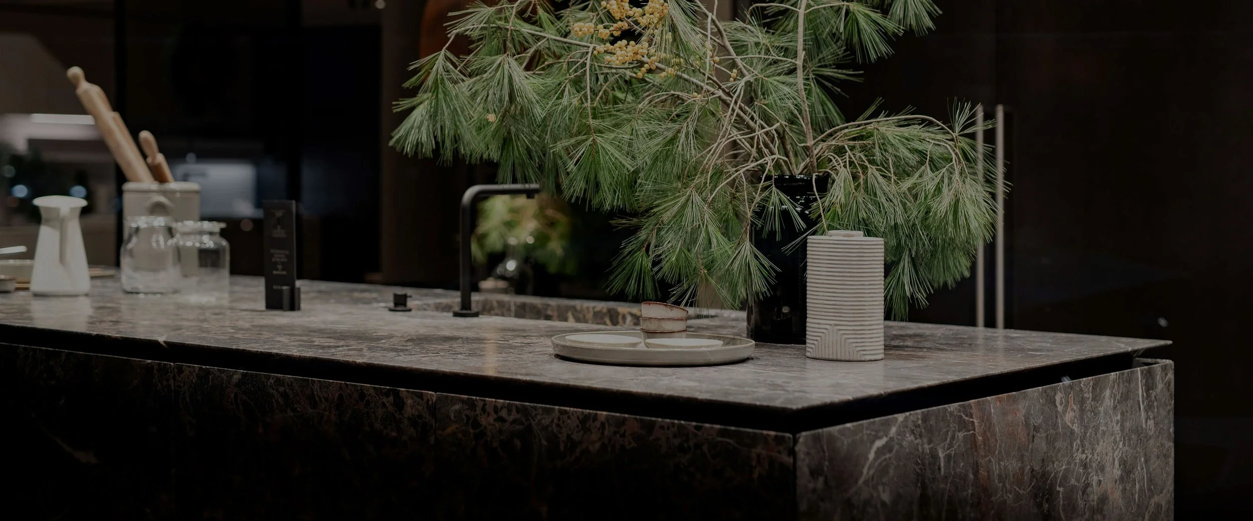 Une cuisine moderne avec un plan de travail en pierre sombre, décorée avec un vase contenant des branches de pin, une assiette avec une tasse, et quelques petits objets.