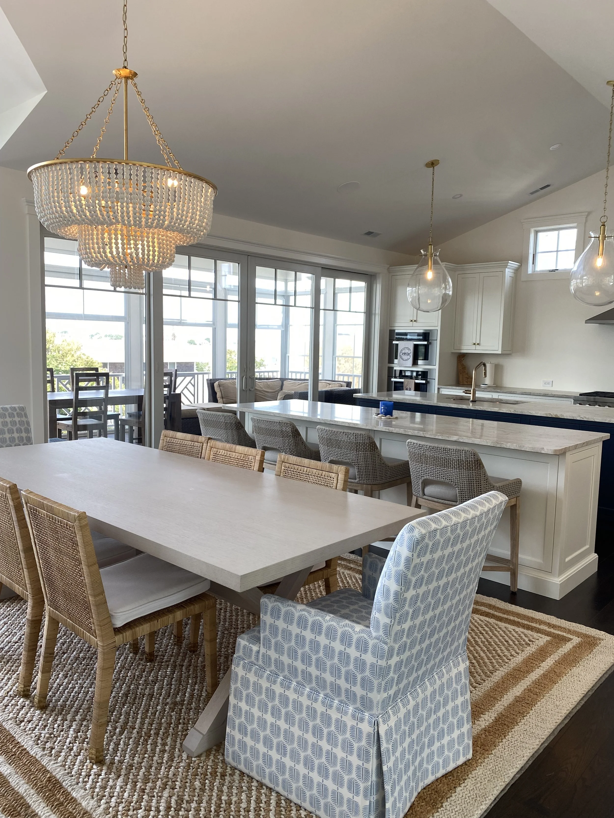 Bright, modern kitchen and dining area with a large wooden table, mixed chairs, a chandelier, and glass pendant lights, overlooking a sunroom with sliding glass doors leading outside.