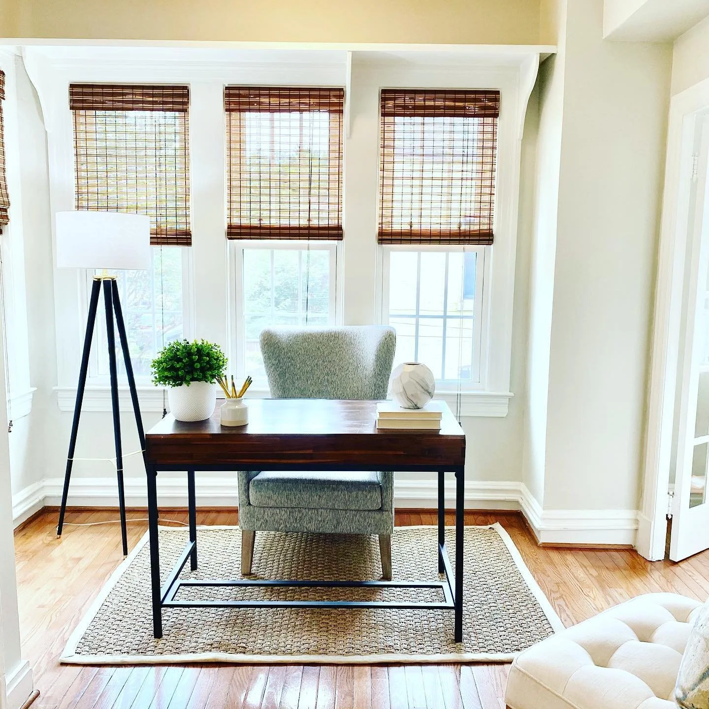 Bright home office with a large window with brown blinds, a desk with a green upholstered chair, a potted plant, and decorative items.