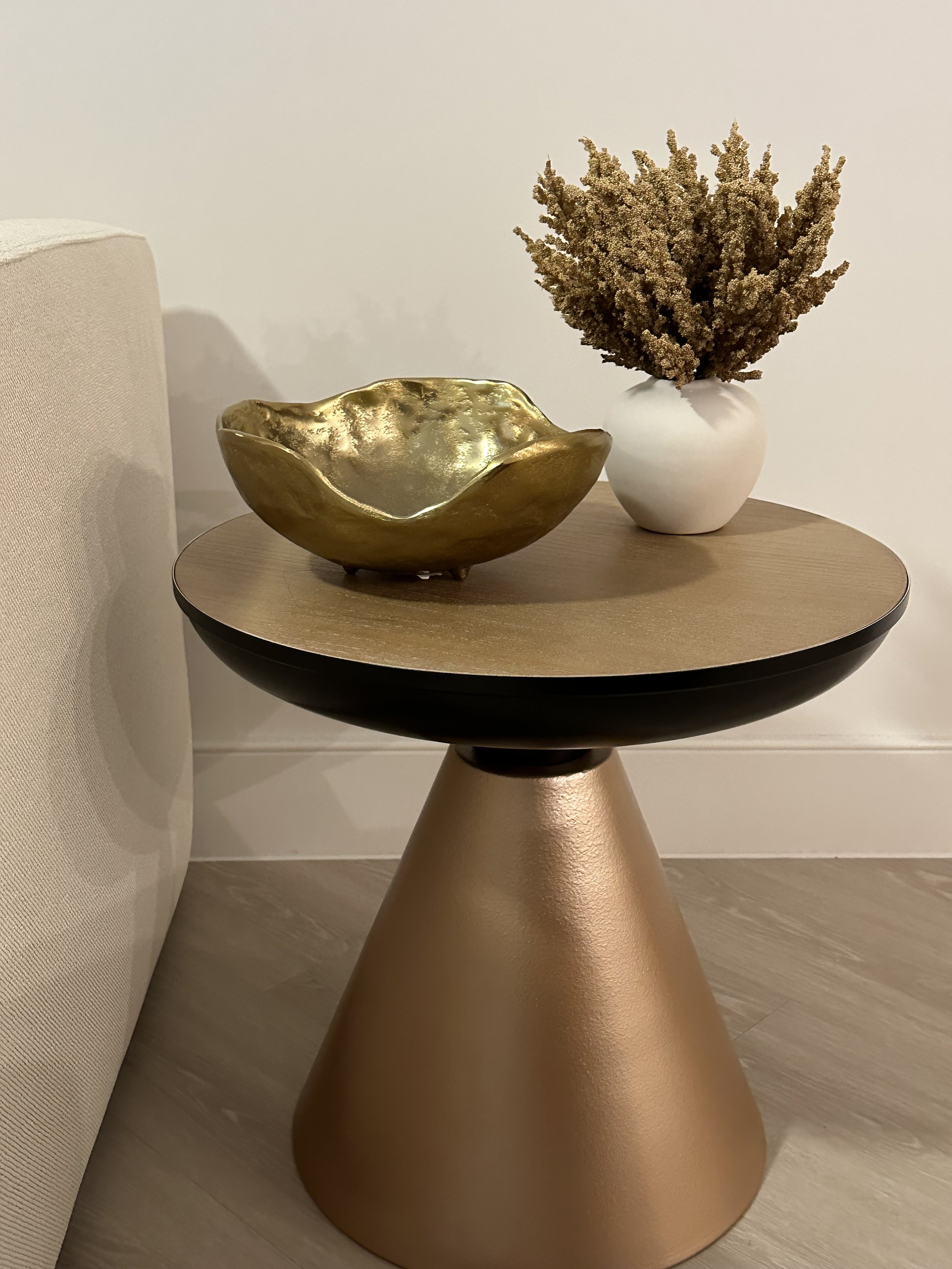 A round side table with a metallic gold base and a wooden top, placed next to a beige sofa. On the table are a gold ceramic bowl and a white vase containing dried flowers.