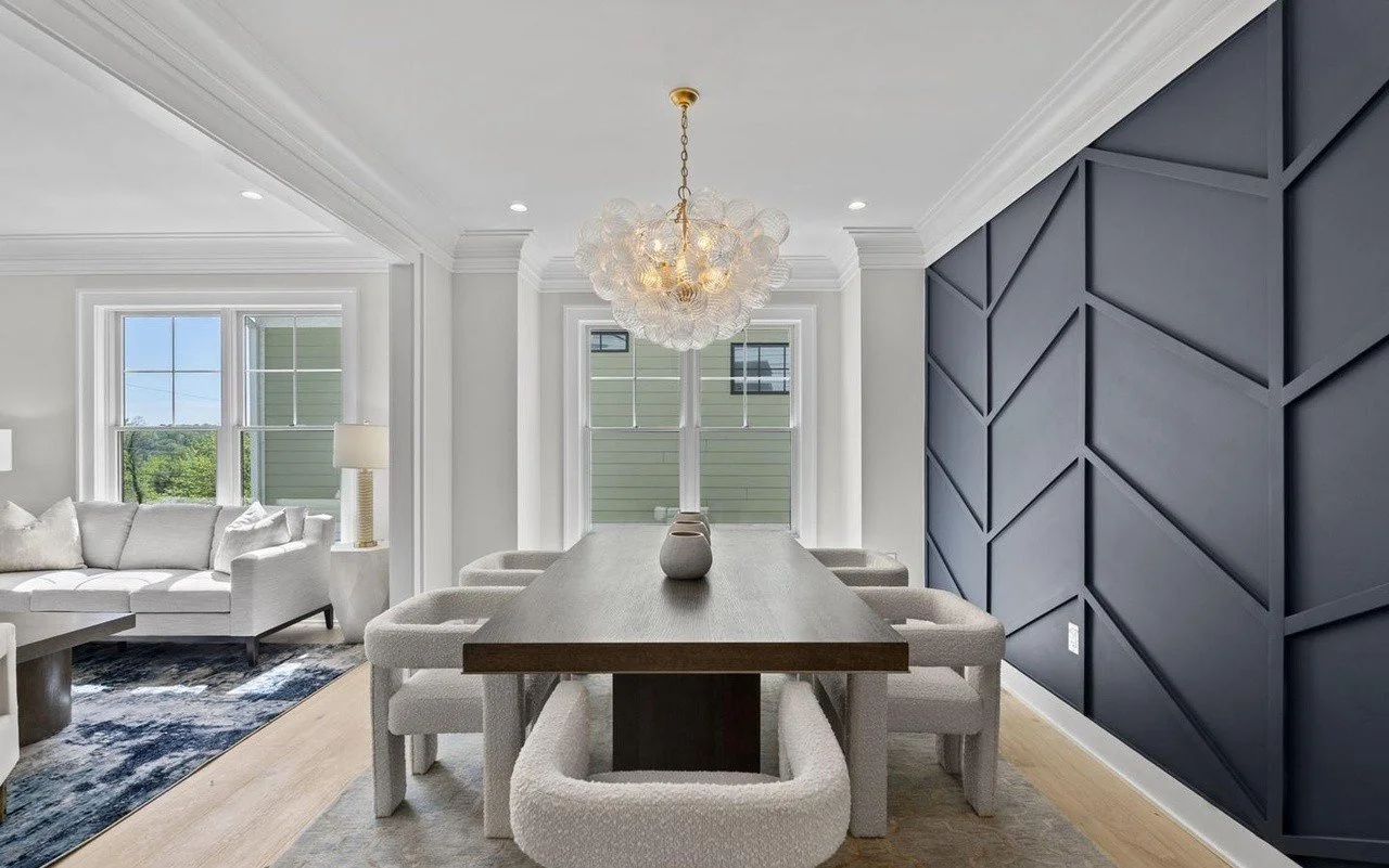 Dining room with a wooden table, six beige upholstered chairs, a modern white vase, a white chandelier, two windows, and a dark blue accent wall with a geometric pattern, adjacent to a living room with a white sofa and a window with a view of green trees.