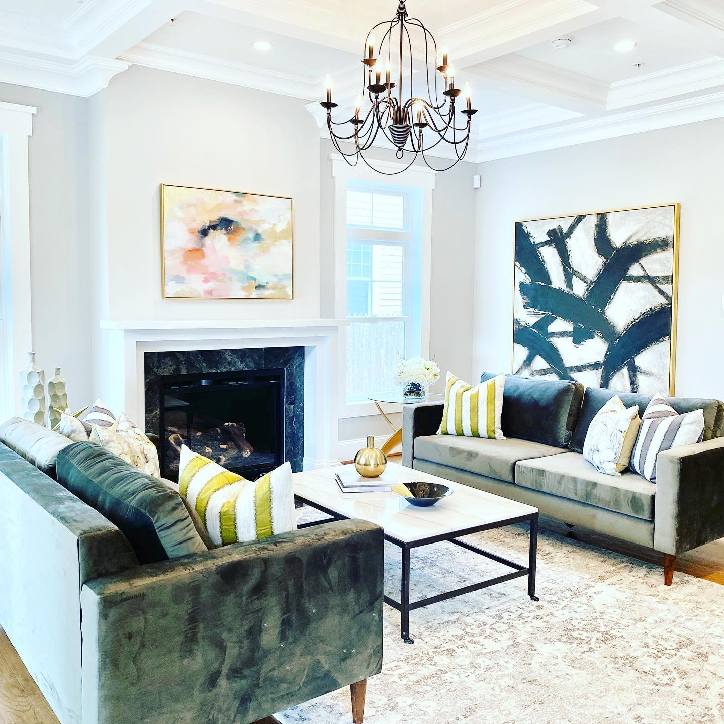 Living room with two sofas, a coffee table, a fireplace, abstract paintings, decorative pillows, a chandelier, and a window with white blinds.