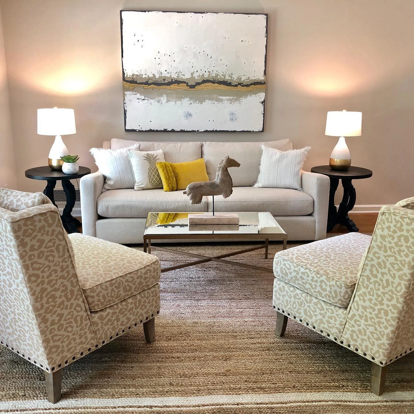 Living room with two patterned armchairs facing each other, a beige sofa with white, yellow, and animal-print pillows, two black side tables with lamps, a wooden horse sculpture on the coffee table, and framed abstract artwork above the sofa.