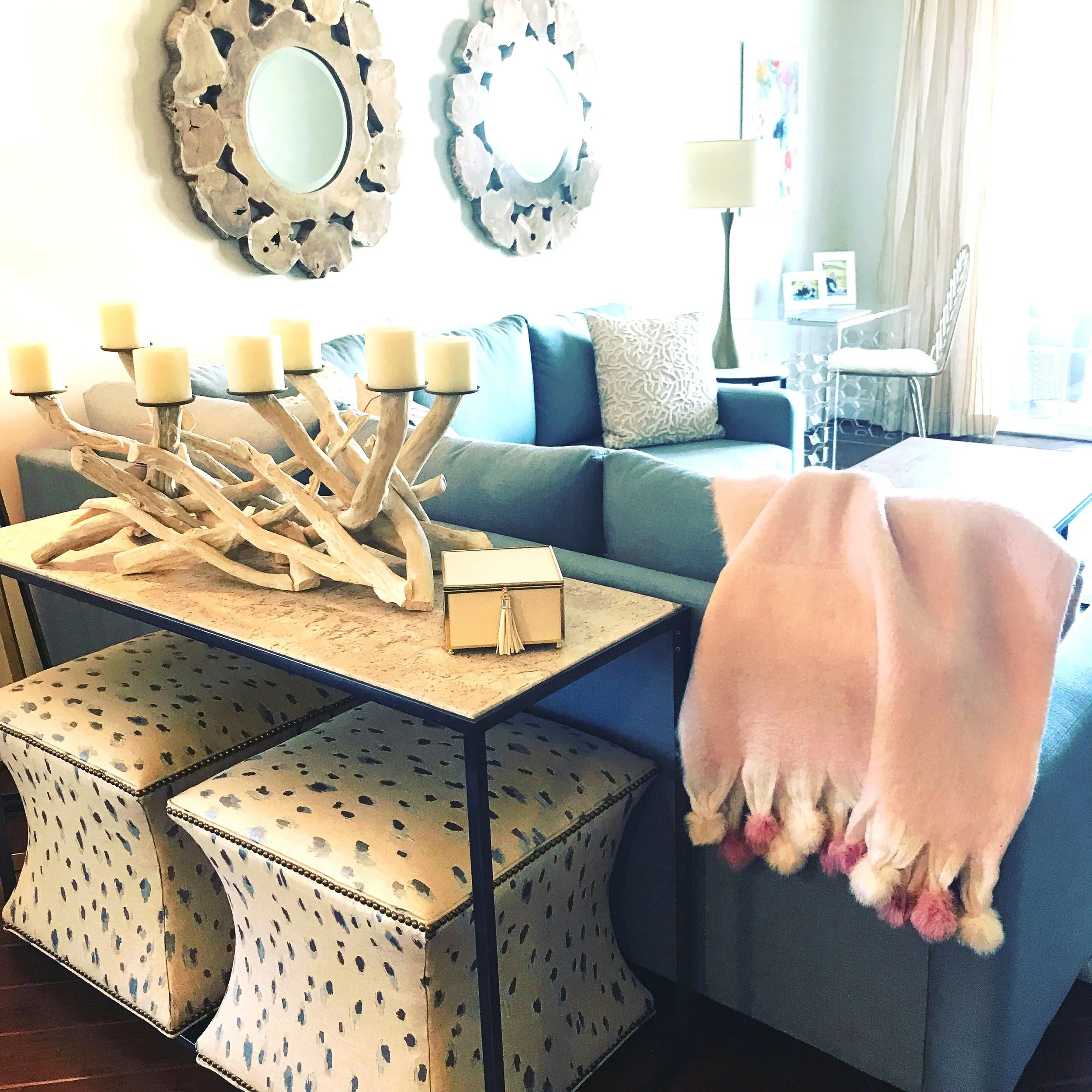 Living room with turquoise sofa, decorative pillows, wall mirrors, standing lamp, table with driftwood candle holder, pink throw blanket with pom-poms, two patterned ottomans, and a workspace with a chair and framed photos.