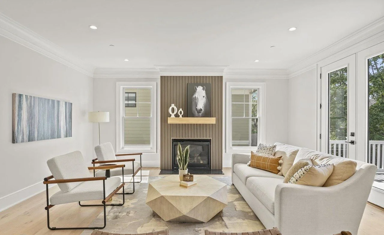 Bright living room with white walls, hardwood floor, modern fireplace with wooden paneling, and large windows revealing trees outside. Furnished with a white sofa, two armchairs with wooden armrests, a geometric coffee table, and a tall floor lamp.