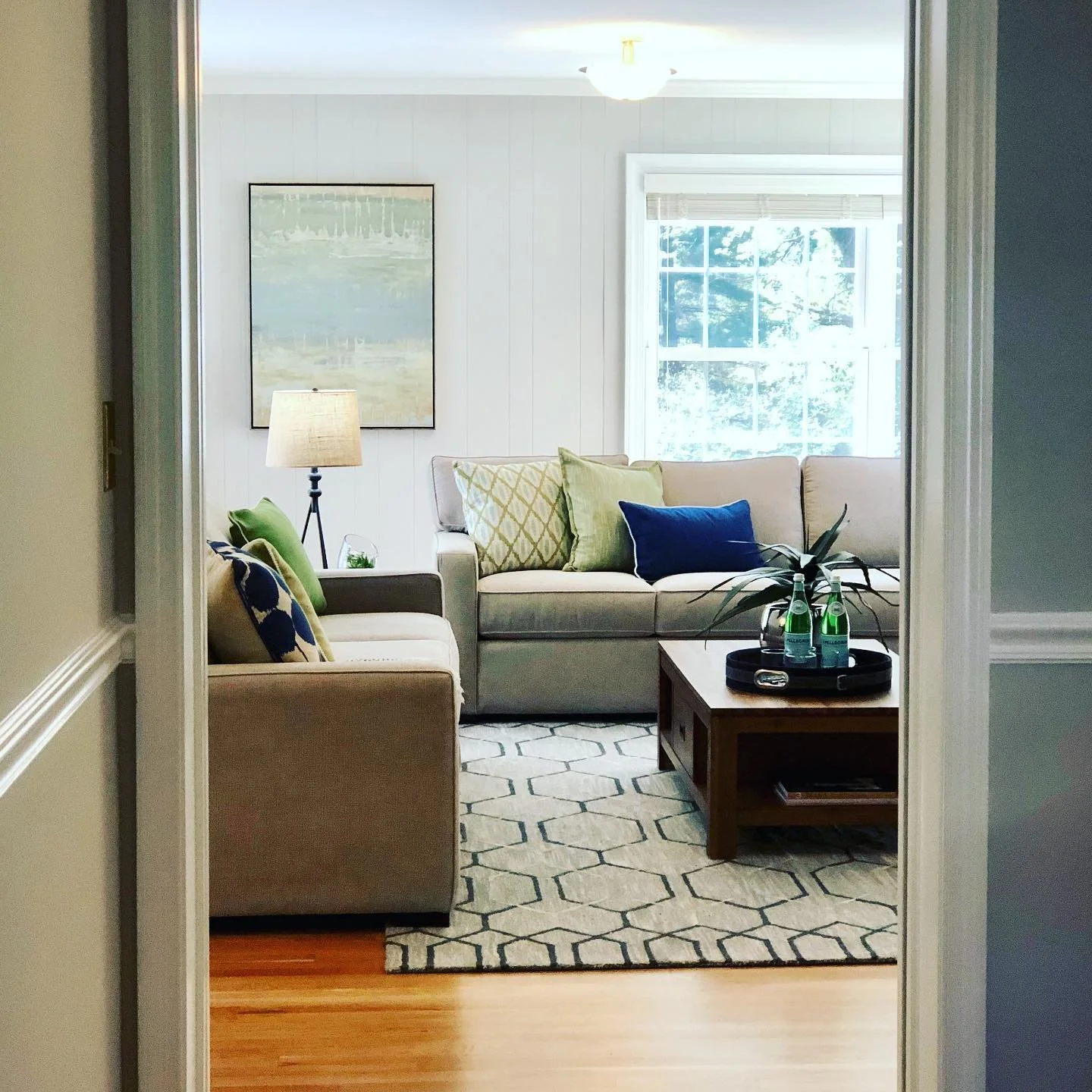 Living room with beige sofa, armchair, wooden coffee table, patterned rug, throw pillows, side lamp, framed art, and window with natural light.