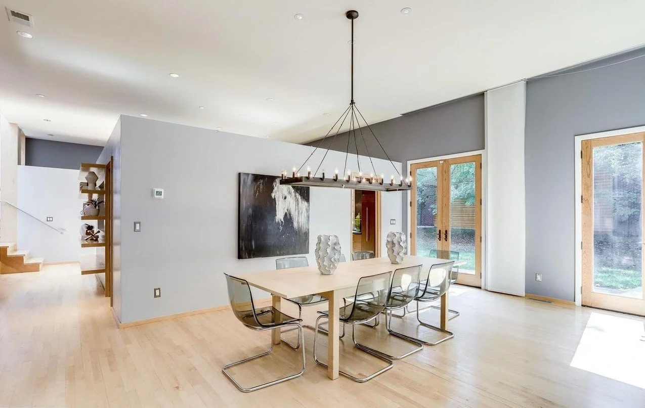 Modern dining room with six transparent chairs around a rectangular light wood table, two white vases, black wall art, large windows, and a chandelier with multiple bulbs.