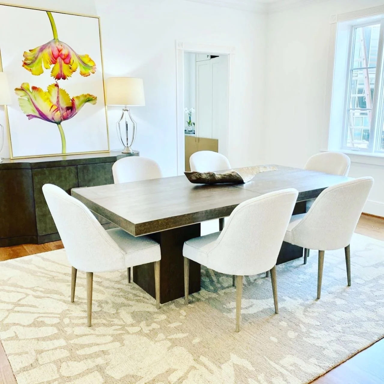 A dining room with a large wooden table surrounded by six white upholstered chairs. There are two table lamps on a sideboard, a colorful floral painting on the wall, and a window letting in natural light.