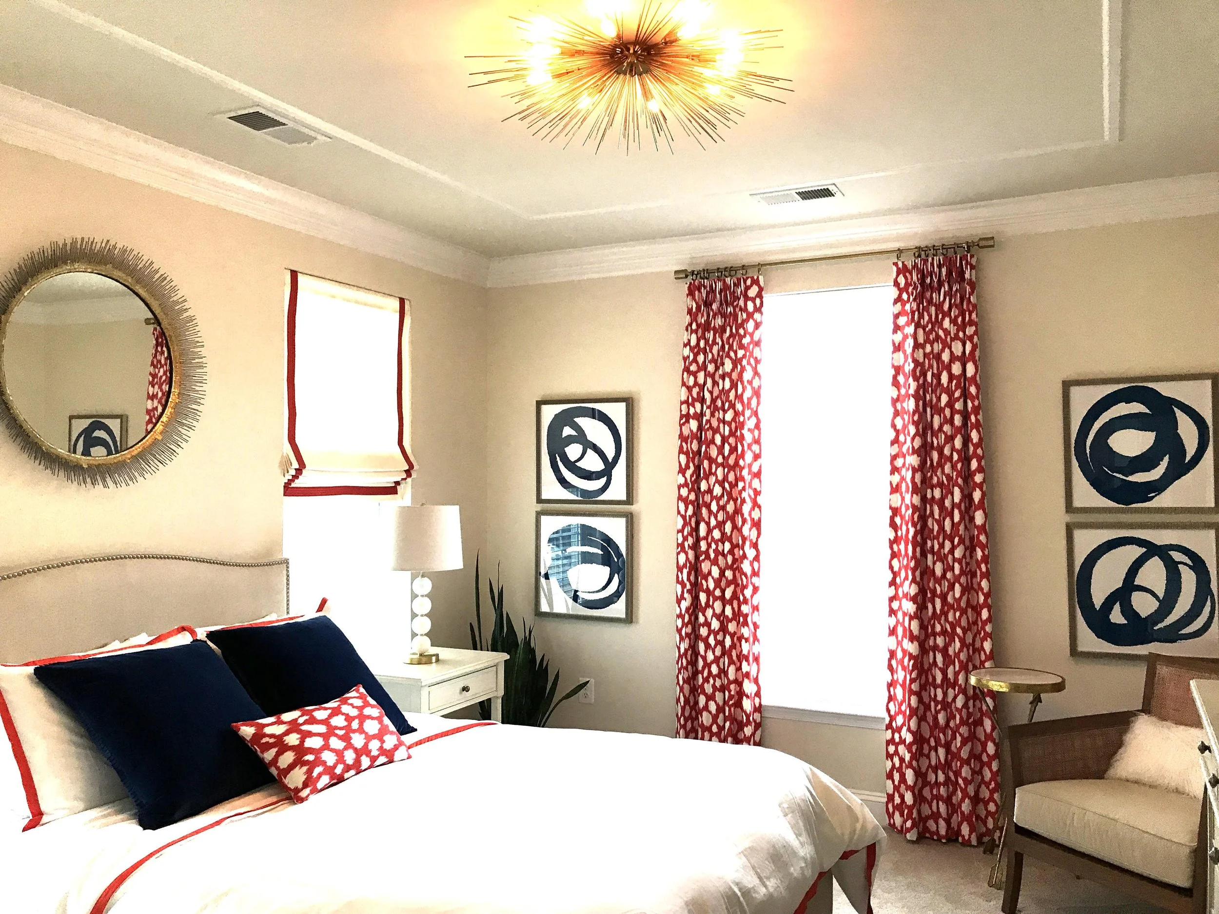 A bedroom with a bed, navy and red pillows, a white bedside table with a white lamp, a mirror on the wall, abstract blue artwork on the walls, a window with patterned red and white curtains, and a beige armchair with a cushion.